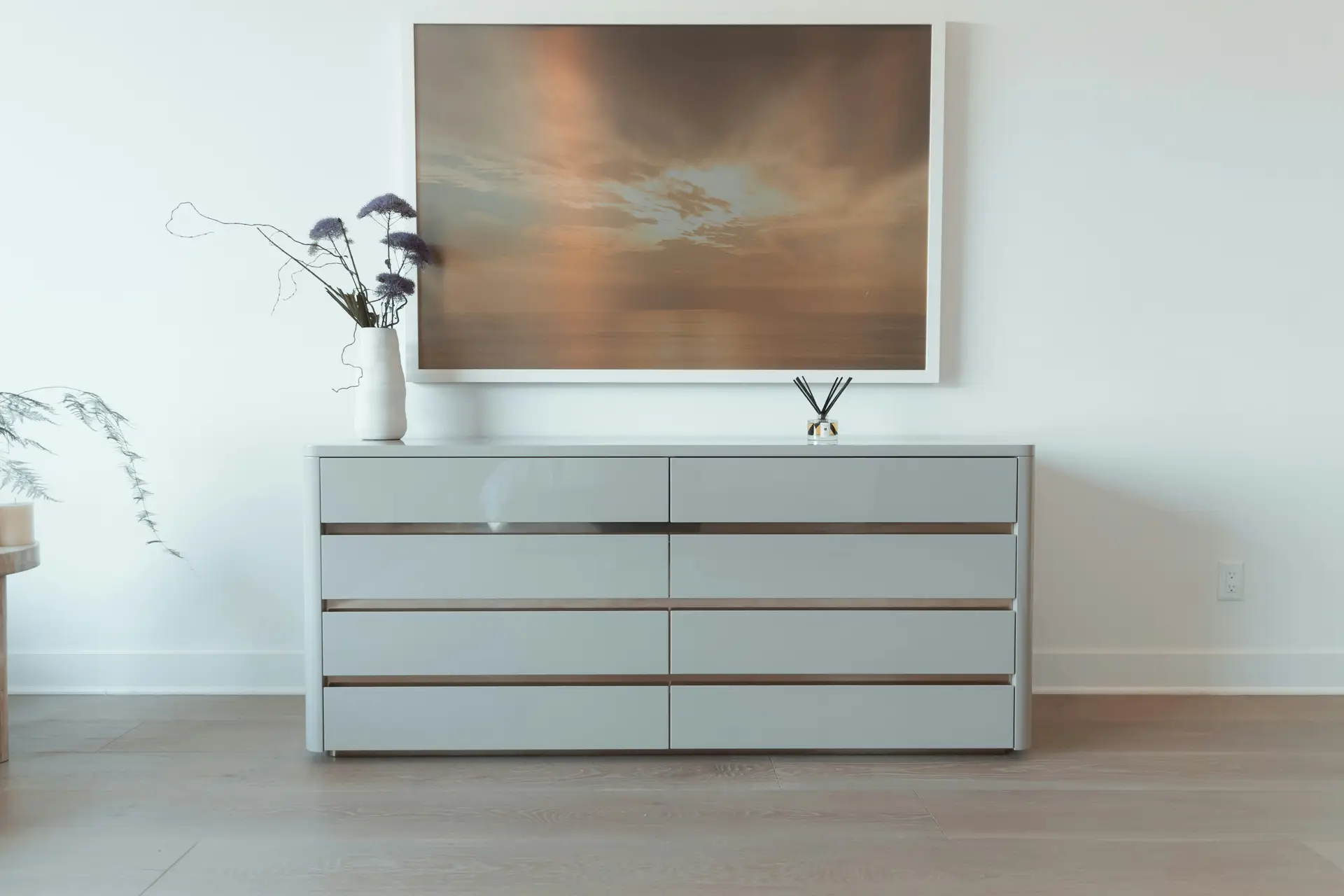 A calm minimal bedroom scene featuring a sleek gray dresser with a decluttered dresser top, styled only with a simple white vase of airy stems and a small reed diffuser. Above it hangs a large, serene landscape artwork, adding a soft, warm focal point against the clean white wall and light wood flooring.
