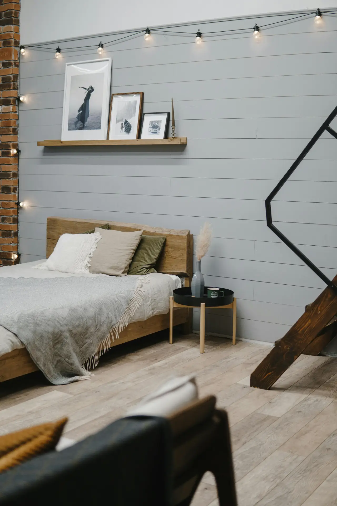 A cozy minimal bedroom with a wooden bed frame, soft layered pillows, and a light woven throw. Above the bed, floating shelves display framed artwork, while string lights add a warm, relaxed glow. A round bedside table with simple decor completes the calm, inviting atmosphere.