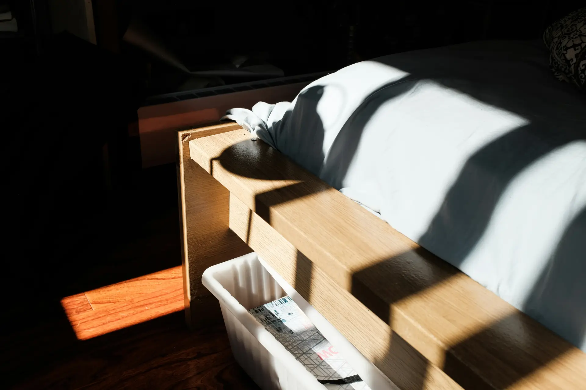 A minimal bedroom scene with morning light casting bold shadows across a wooden hidden storage bed, revealing a discreet bin tucked underneath and soft bedding draped loosely over the frame for a clean, understated look.