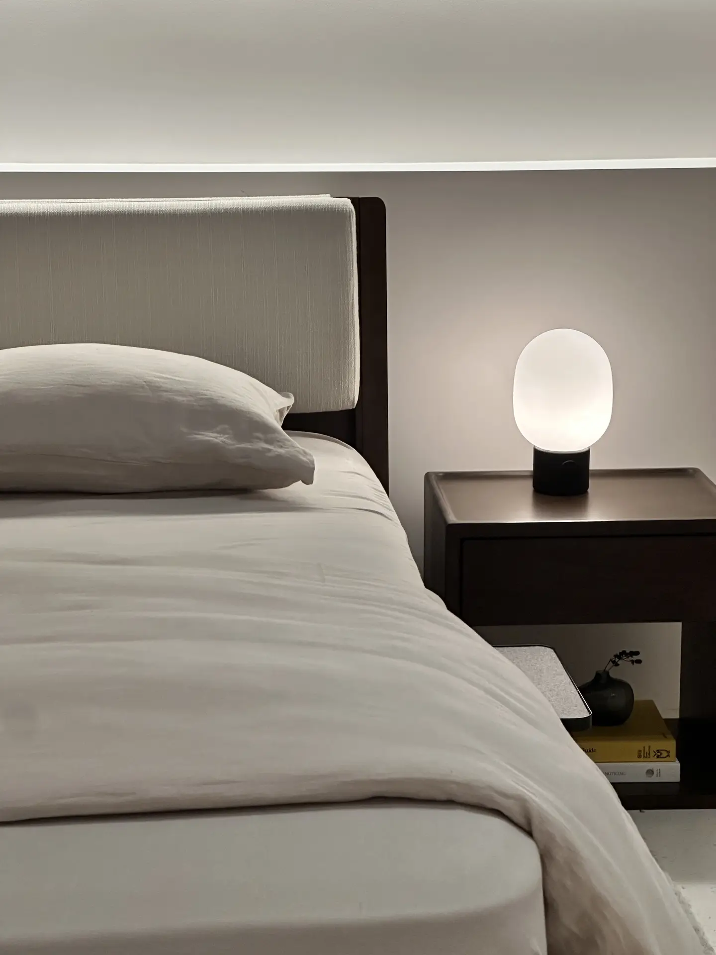 A serene minimal bedroom featuring soft neutral bedding, a simple upholstered headboard, and minimal nightstands in a dark wood tone. A rounded, glowing table lamp adds gentle ambient light, while a few neatly arranged books and a small decorative accent keep the space clean and versatile without disrupting the calm aesthetic.