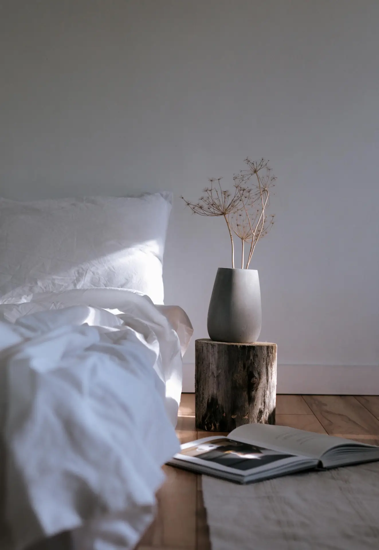 A serene minimal bedroom with sunlight hitting crisp white bedding, a simple vase of dried stems resting on a natural wood stump used as a side table. An open book lies on the floor nearby, enhancing the relaxed, effortless atmosphere of the space.