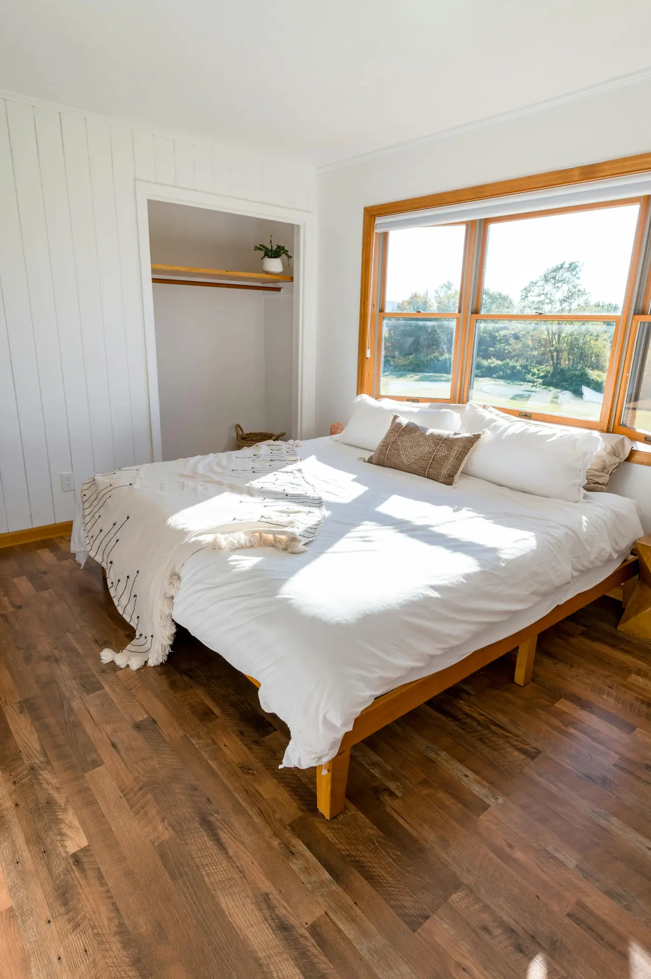 A bright minimal bedroom with sunlight streaming across a simple wooden bed dressed in white bedding and soft neutral pillows. The open closet and uncluttered floor highlight a sense of negative space appreciation, allowing the warm wood tones and serene outdoor view to stand out without distraction.