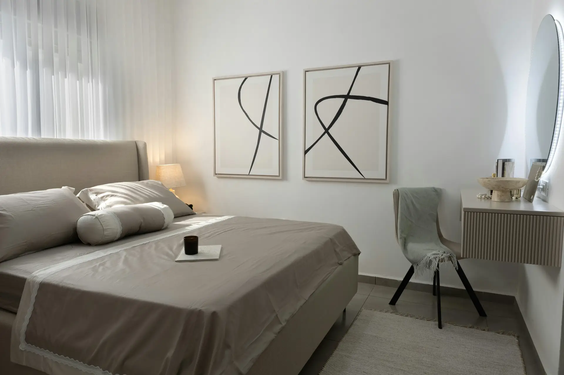 A bright minimal bedroom with neutral textured bedding, soft beige tones, and clean-lined furniture. Two abstract black-and-white art pieces hang above the bed, while a floating vanity with a sculptural bowl and mirror adds a refined touch. Light curtains, a simple chair draped with a throw, and a small bedside lamp create an inviting, versatile atmosphere.