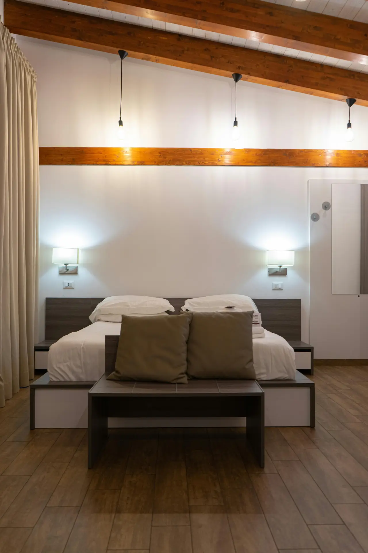 A warm minimal bedroom with wooden beams overhead, soft lighting, and a neatly made bed framed by matching wall sconces. A bench with neutral cushions sits at the foot of the bed, and the entire setup looks clean and intentional with no visible cables, giving the space a tidy, streamlined feel.