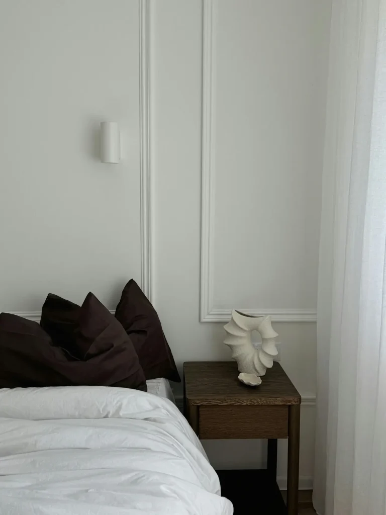 A refined minimal bedroom with crisp white bedding, dark pillows for contrast, and soft natural light filtering through sheer curtains. A wooden nightstand holds one statement piece-a sculptural ceramic vase-set against a wall with subtle panel detailing and a compact white sconce, creating a clean yet expressive look.