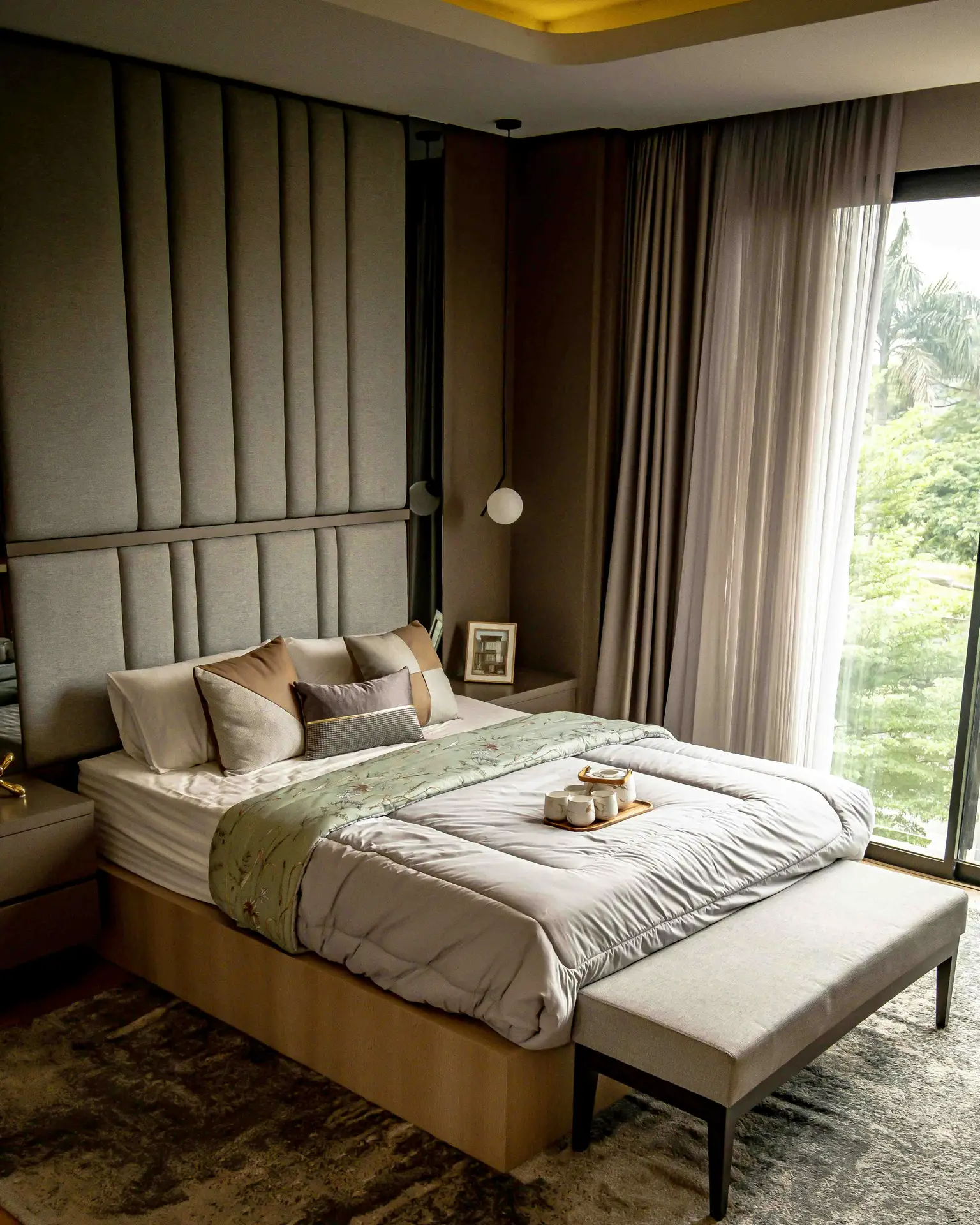 A warm minimal bedroom with layered neutral bedding, a tall upholstered headboard, and a bench at the foot of the bed. Soft sheer curtains filter natural light from the large window, while hanging globe lights and a few simple accents create a calm, elevated atmosphere.