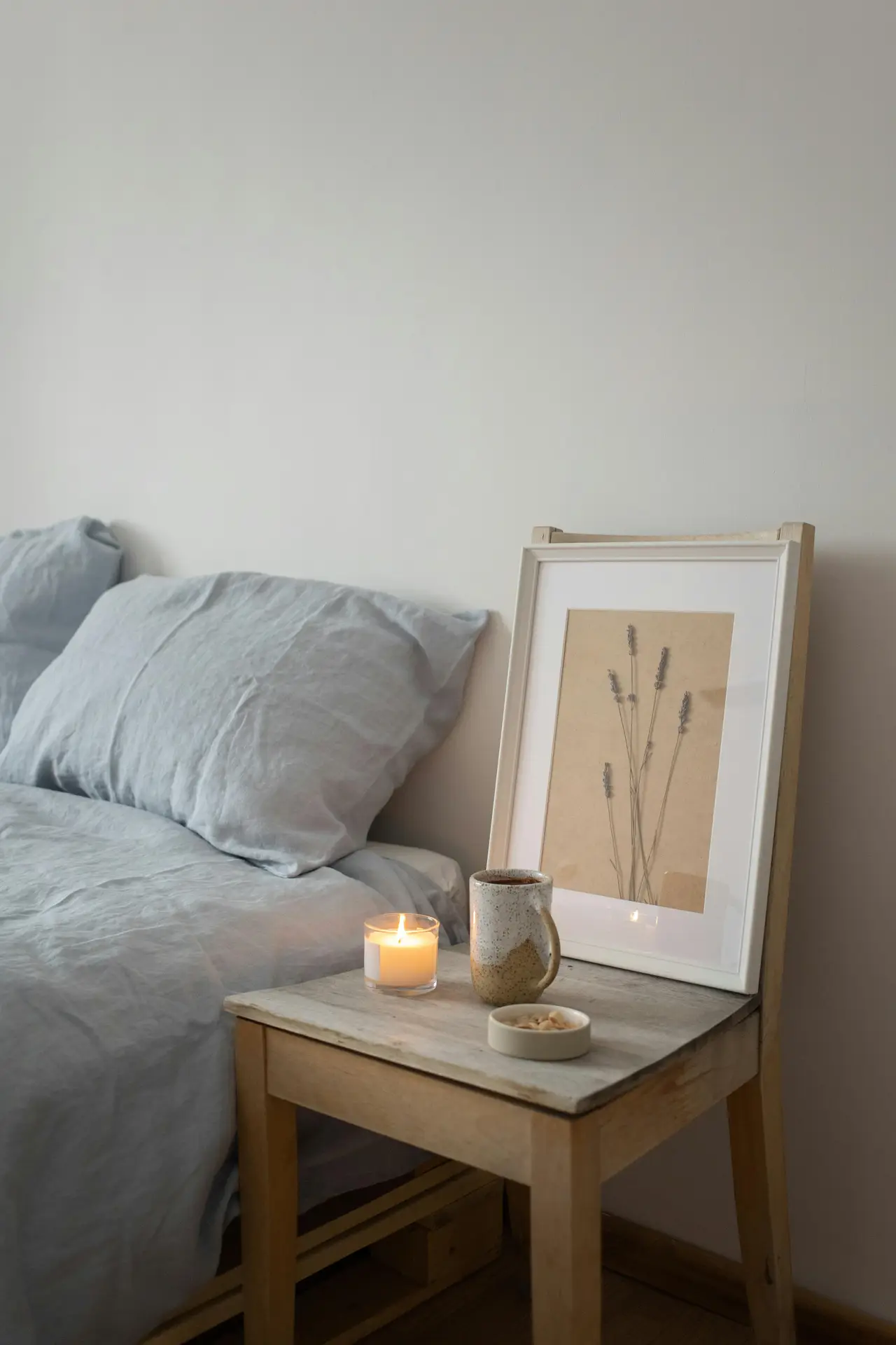 A peaceful minimal bedroom with muted bedding and a simple wooden chair used as a bedside table. A lit candle adds soft scents to the space, accompanied by a ceramic mug and framed botanical print, creating a calm, cozy corner with a natural, understated charm.