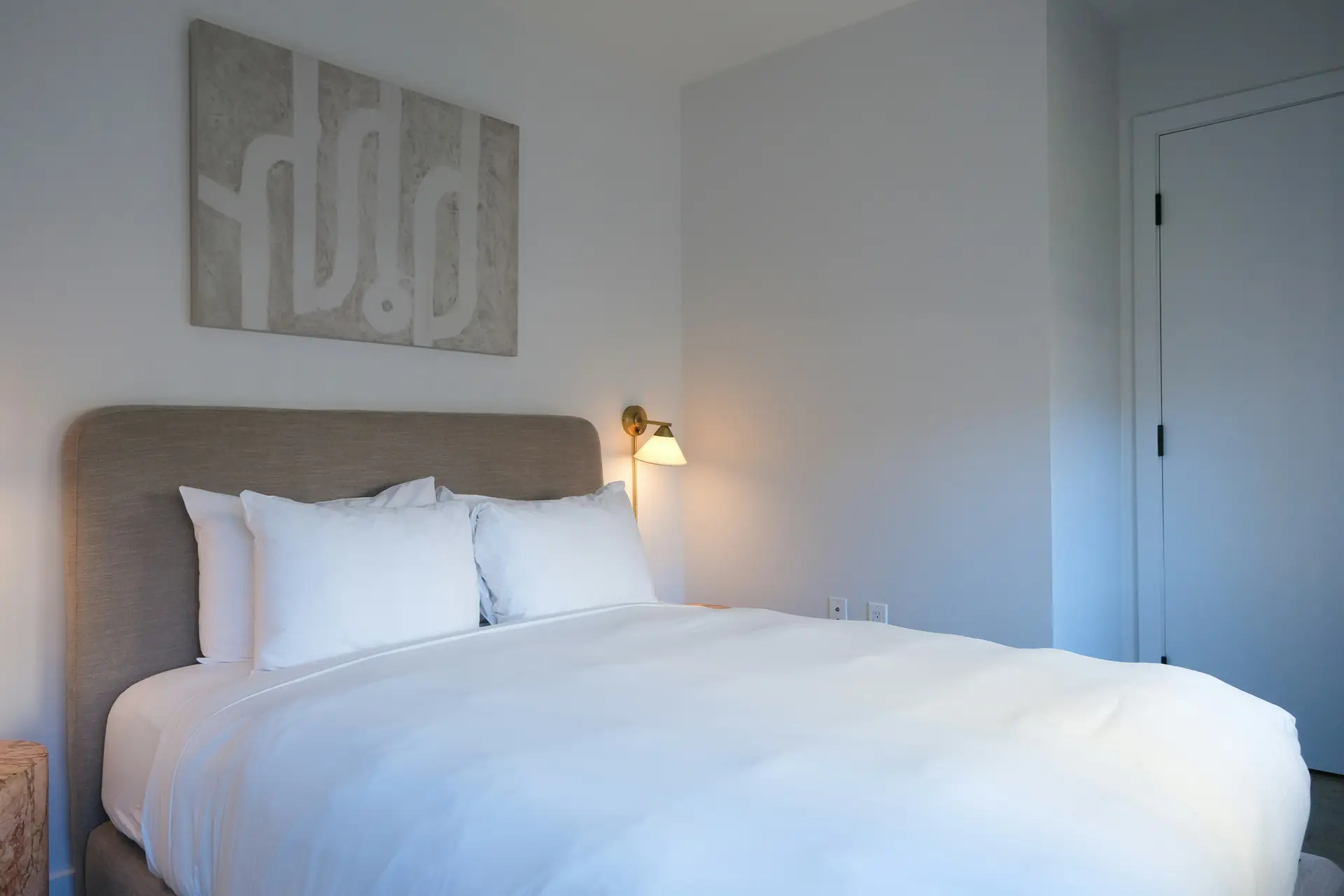 A calm minimal bedroom with a soft upholstered headboard, crisp white bedding, and warm bedside lighting. Above the bed hangs a tone-in-tone artwork that blends seamlessly with the muted wall color, adding subtle texture without overwhelming the clean, serene look of the space.