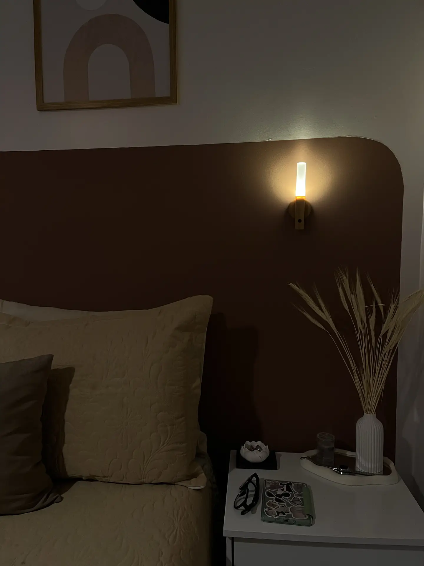 A cozy minimal bedroom featuring warm-toned bedding, a curved painted accent wall, and soft wall-mounted lighting that casts a gentle glow. A small nightstand holds simple decor like a vase with dried stems, a tray with essentials, and a pair of glasses, creating a calm and practical nighttime setup.