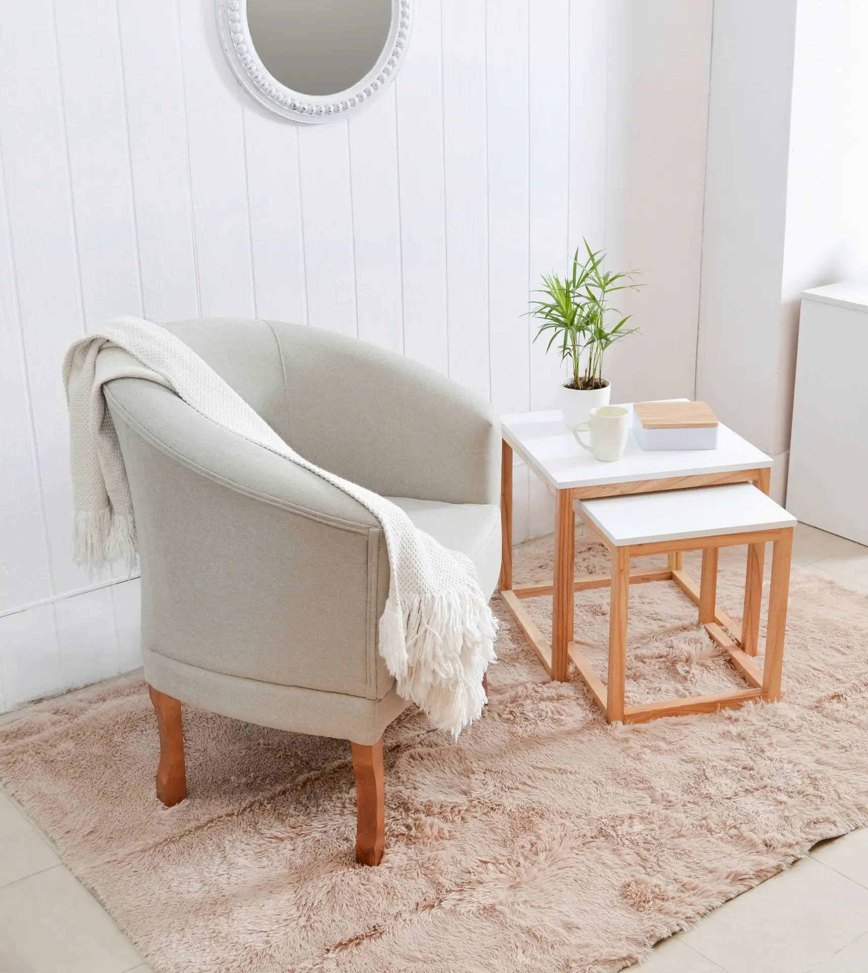 A cozy minimalist living room corner featuring a beige armchair with a soft throw, a pair of white nesting tables with wooden legs, and a fluffy neutral rug. The mix of natural wood, fabric, and light tones creates a sense of harmony and cohesive materials throughout the space.