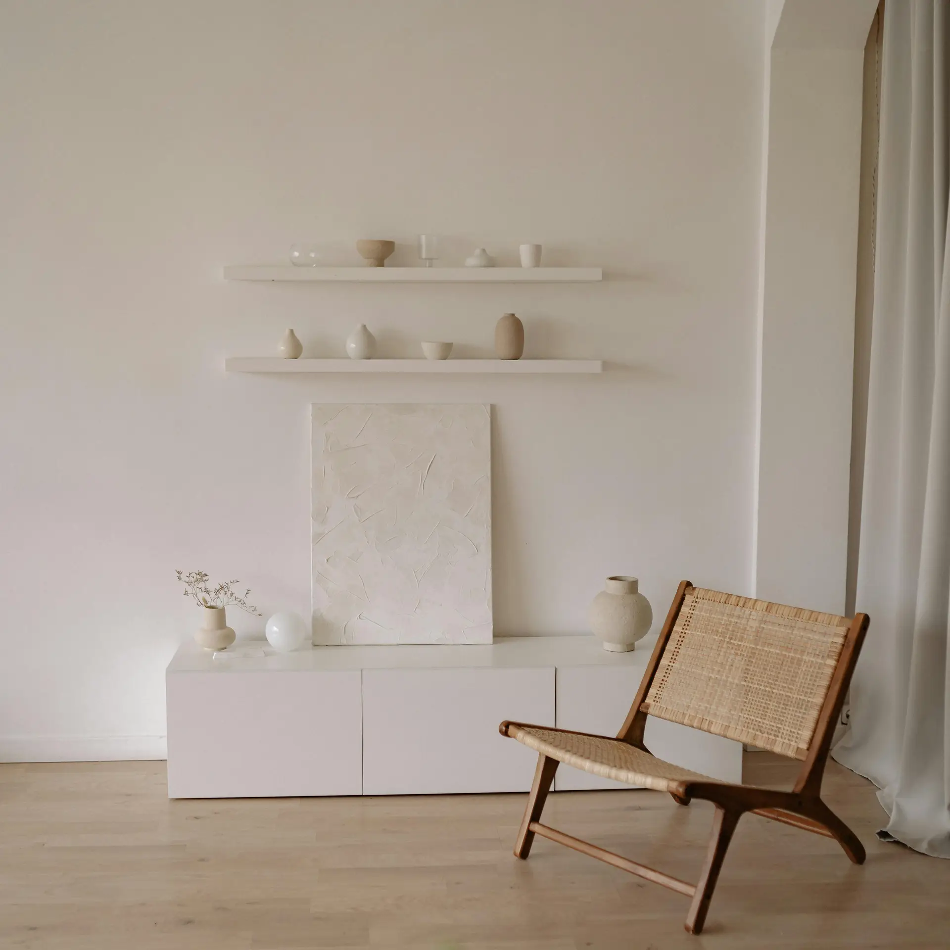 A minimalist living room with floating furniture, featuring sleek white shelves and a wall-mounted console decorated with neutral vases and ceramics. A woven chair in natural wood tones adds warmth and texture, balancing the clean and airy aesthetic of the space.