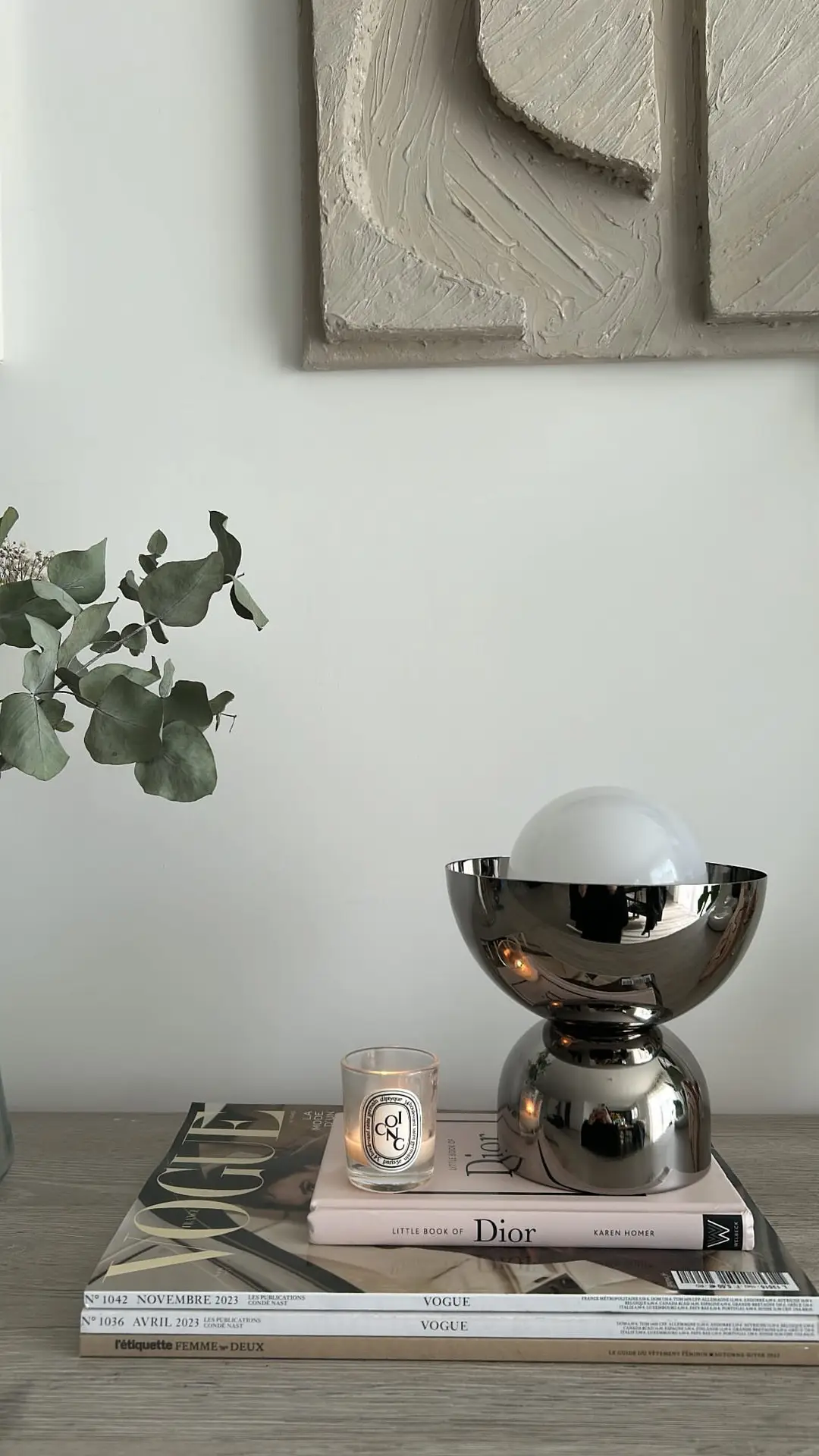 A minimalist living room setup featuring a sleek metallic lamp, a lit candle, and a small stack of fashion books arranged neatly on a wooden surface. The muted color palette and sculptural wall art above highlight a balance between functionality and decorative appeal, creating an elegant example of functional decor.