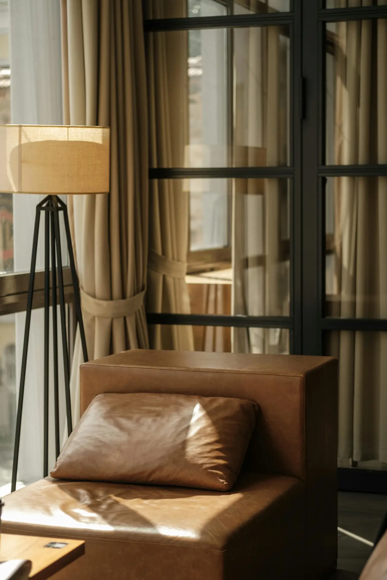 A minimalist living room featuring a brown leather armchair positioned beside a modern floor lamp with a linen shade. Natural sunlight filters through large windows with layered curtains, creating a warm and inviting lighting composition.