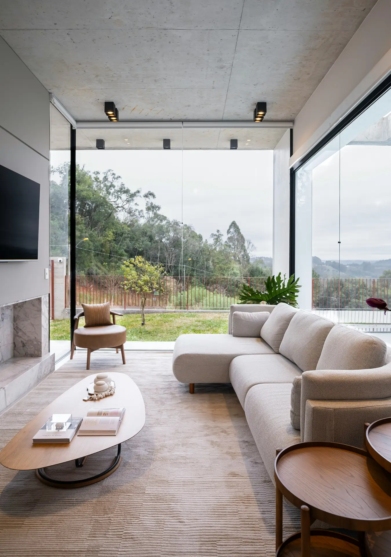A bright minimalist living room with floor-to-ceiling windows overlooking a green landscape, featuring a neutral-toned sectional sofa, a sleek coffee table, and a single armchair. The open layout and uncluttered arrangement highlight a refined sense of calm and negative space appreciation.