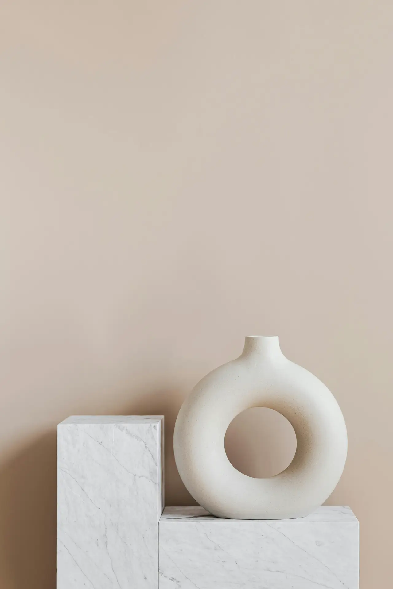 A minimalist living room detail showcasing a circular sculptural vase with a central opening placed on a marble pedestal. The soft beige background and clean lines highlight the refined elegance and focus on sculptural decor pieces within a serene, modern setting.