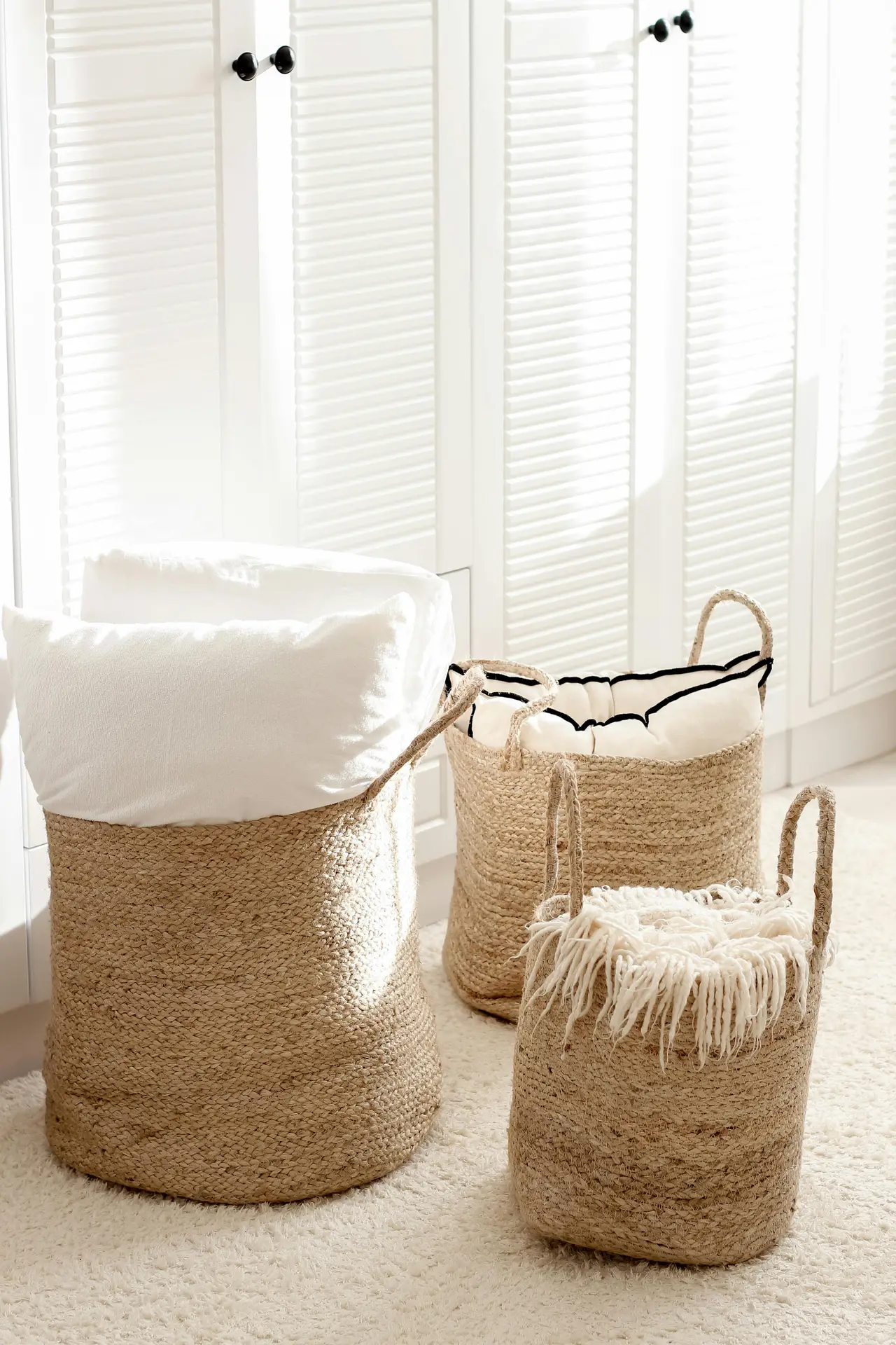 A minimalist living room corner featuring three woven baskets used as smart storage solutions. The baskets hold soft pillows and throws, blending natural textures with a neutral palette that adds warmth and functionality to the bright, airy space.