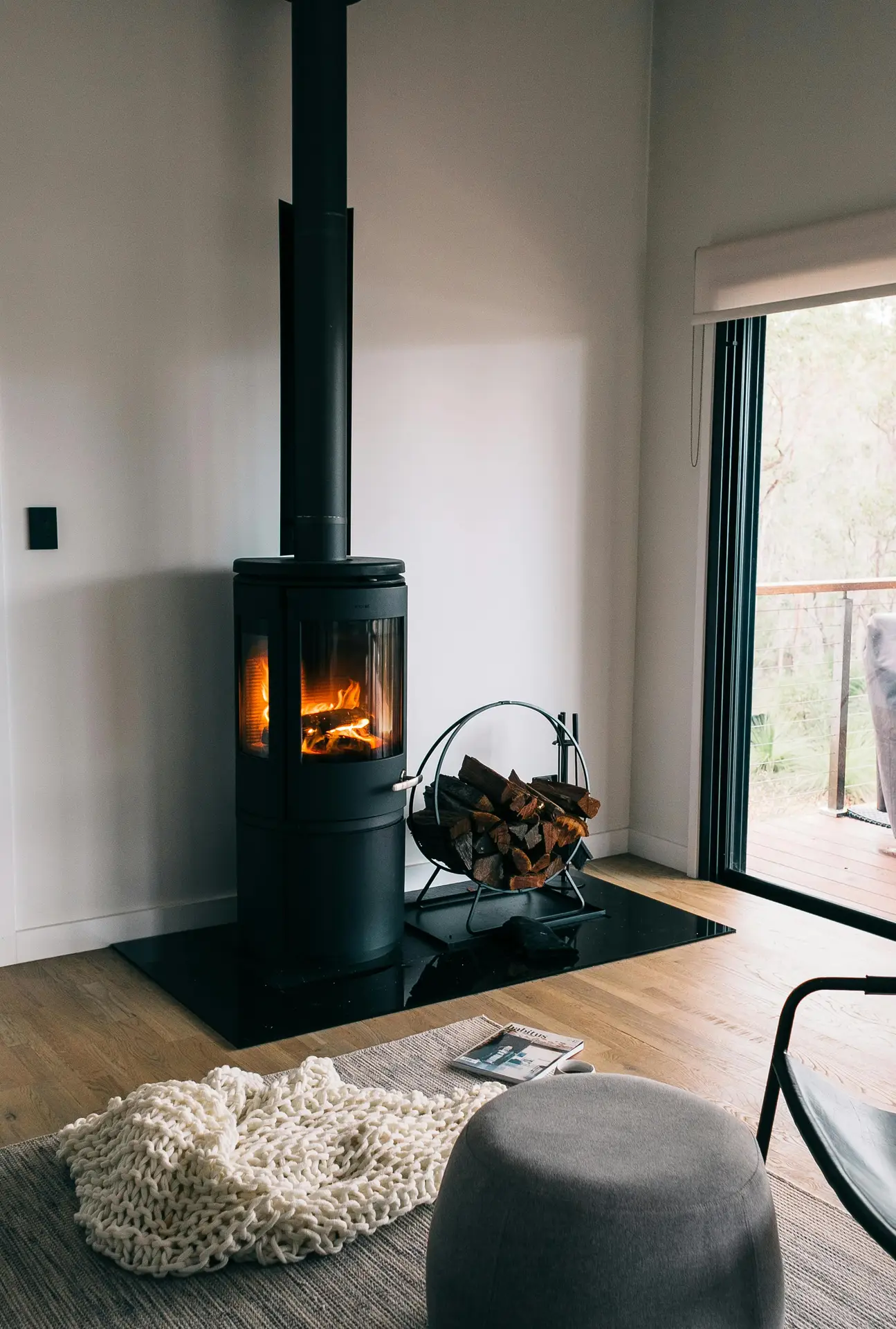 A cozy Nordic living room featuring soft wood floors, a modern black wood-burning stove, and matching black accents like the log holder and sliding door frame. A chunky knit throw, a neutral pouf, and a simple rug create a warm, relaxed atmosphere next to the glowing fire.