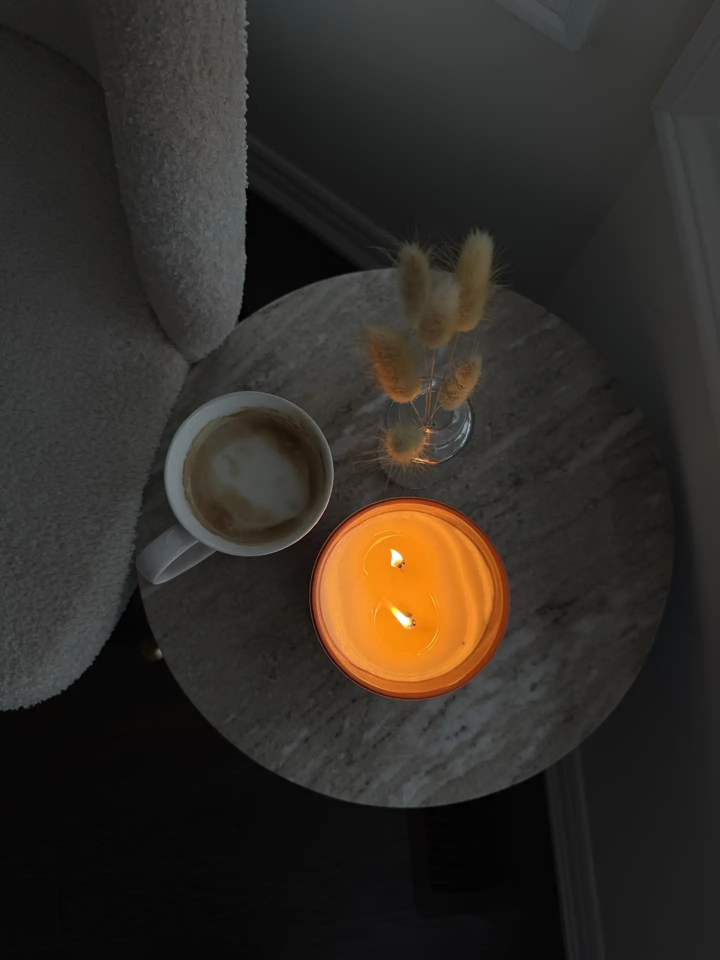 A warm Nordic living room moment with candles and ambient light, showing a glowing orange candle on a round stone side table beside a half-finished coffee and a small vase of dried stems. Soft shadows and a textured chair edge create a calm, intimate atmosphere.