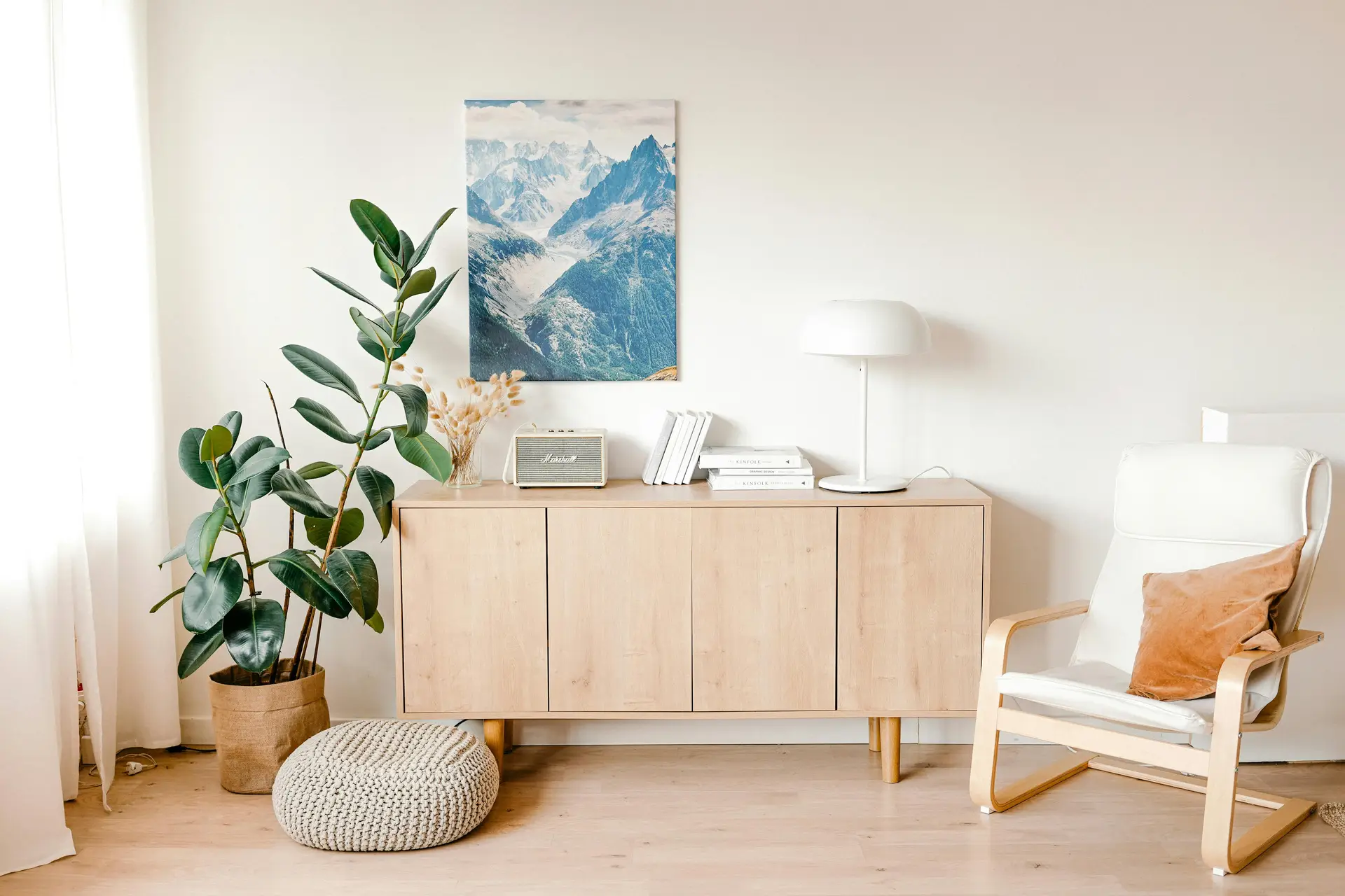 A bright Nordic living room featuring a functional sideboard in light wood, styled with a table lamp, stacked books, dried florals, and a small speaker. A large plant and a knitted floor pouf add warmth, while a simple armchair with a soft cushion completes the airy, minimal setup.