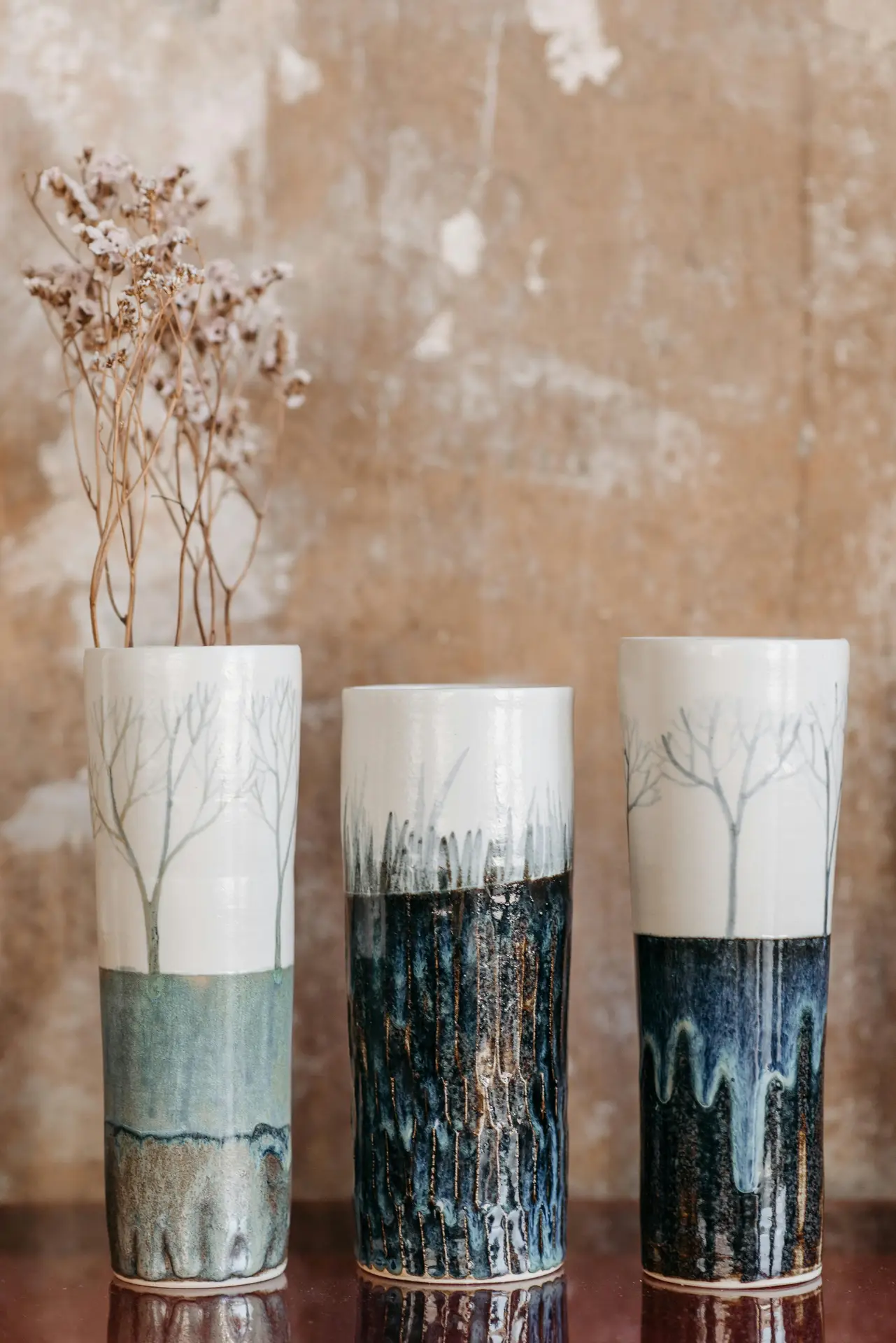 A set of tall ceramic vases with nature-inspired glazing sits against a textured wall, adding an artisanal touch that fits effortlessly into a Nordic living room. The hand crafted decor features soft tree motifs and earthy blue tones, paired with dried stems for a calm, organic look.