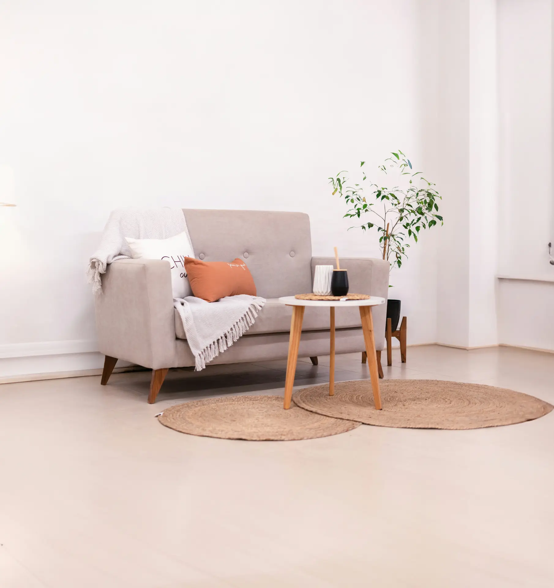 A bright Nordic living room styled with layered neutrals, featuring a light beige loveseat with soft throws and pillows, a round wooden side table, and two woven jute rugs on a pale floor. A simple potted plant in the corner and minimal accessories create a calm, airy Scandinavian feel.