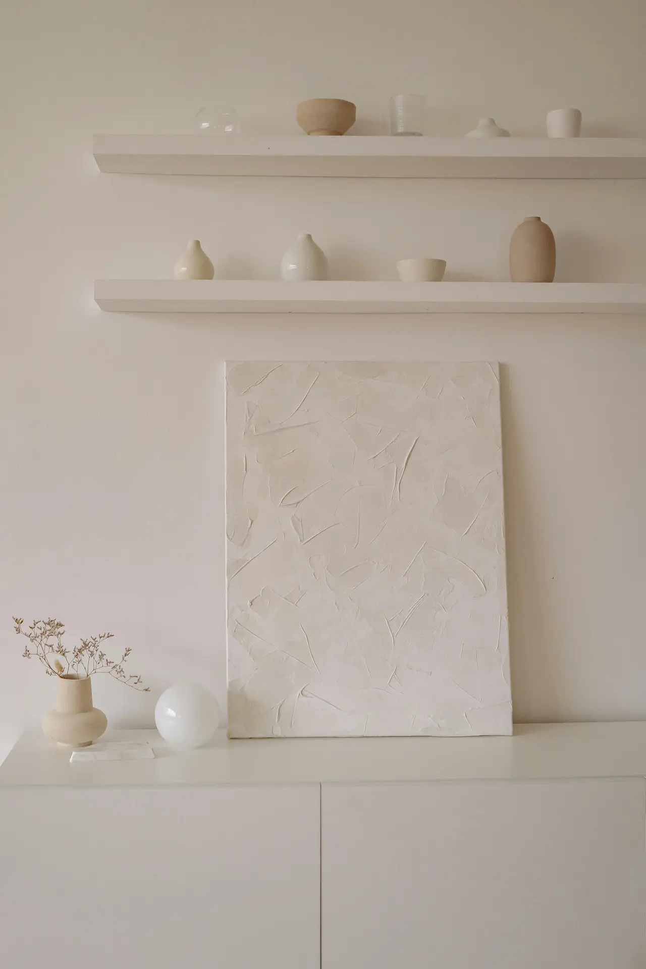 A soft, neutral-toned scene in a Nordic living room featuring minimal wall art, with a textured white canvas leaning against a cabinet and two floating shelves above it holding small ceramic pieces and glass decor.