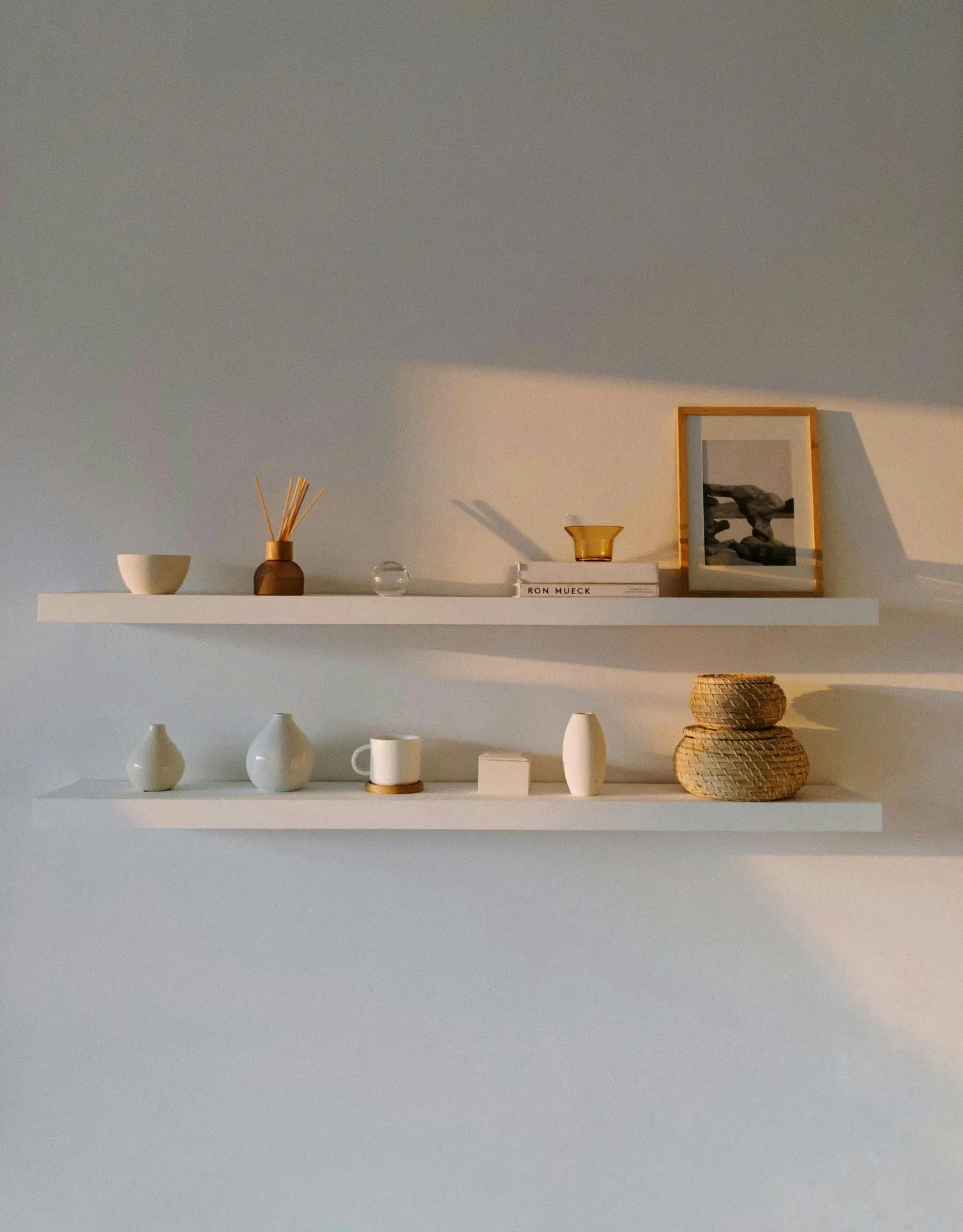 A Nordic living room setup with two open shelves styled in soft evening light, holding simple ceramics, woven baskets, a framed photo, and a few books. The clean arrangement and warm shadows create a calm, minimalist atmosphere.