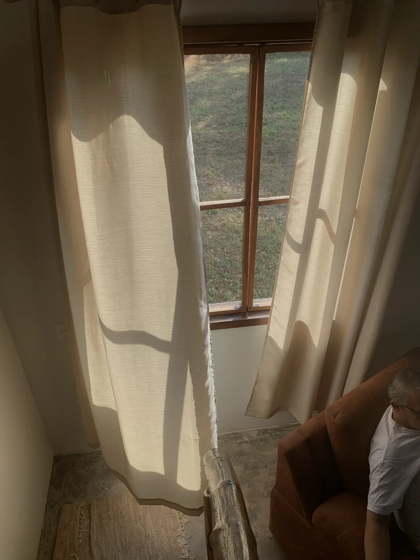 A warm Nordic living room scene with soft linen curtains catching the afternoon light, casting gentle shadows across the floor. A wooden-framed window opens to a grassy view outside, while a muted-toned armchair sits in the corner, creating a calm and understated atmosphere.