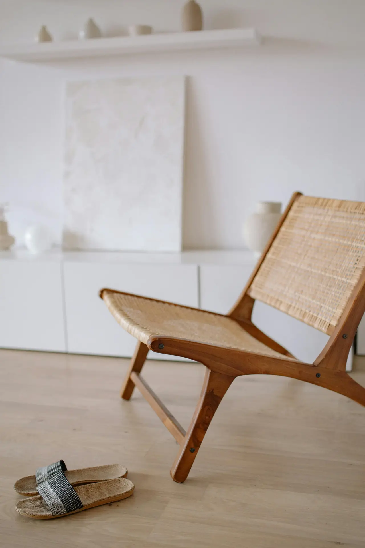 A calm Nordic living room scene featuring a woven wooden statement chair set on light flooring, with simple white cabinetry and soft-toned decor in the background. A pair of minimalist slippers rests nearby, adding to the relaxed, airy atmosphere.