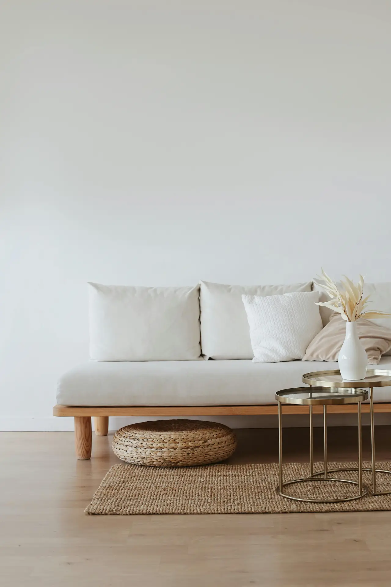 A serene Nordic living room featuring a low, light-wood sofa with soft neutral cushions, paired with gold nesting tables holding a simple white vase. A woven textured rug and a round natural floor cushion add warmth and subtle detail to the minimalist, airy setup.
