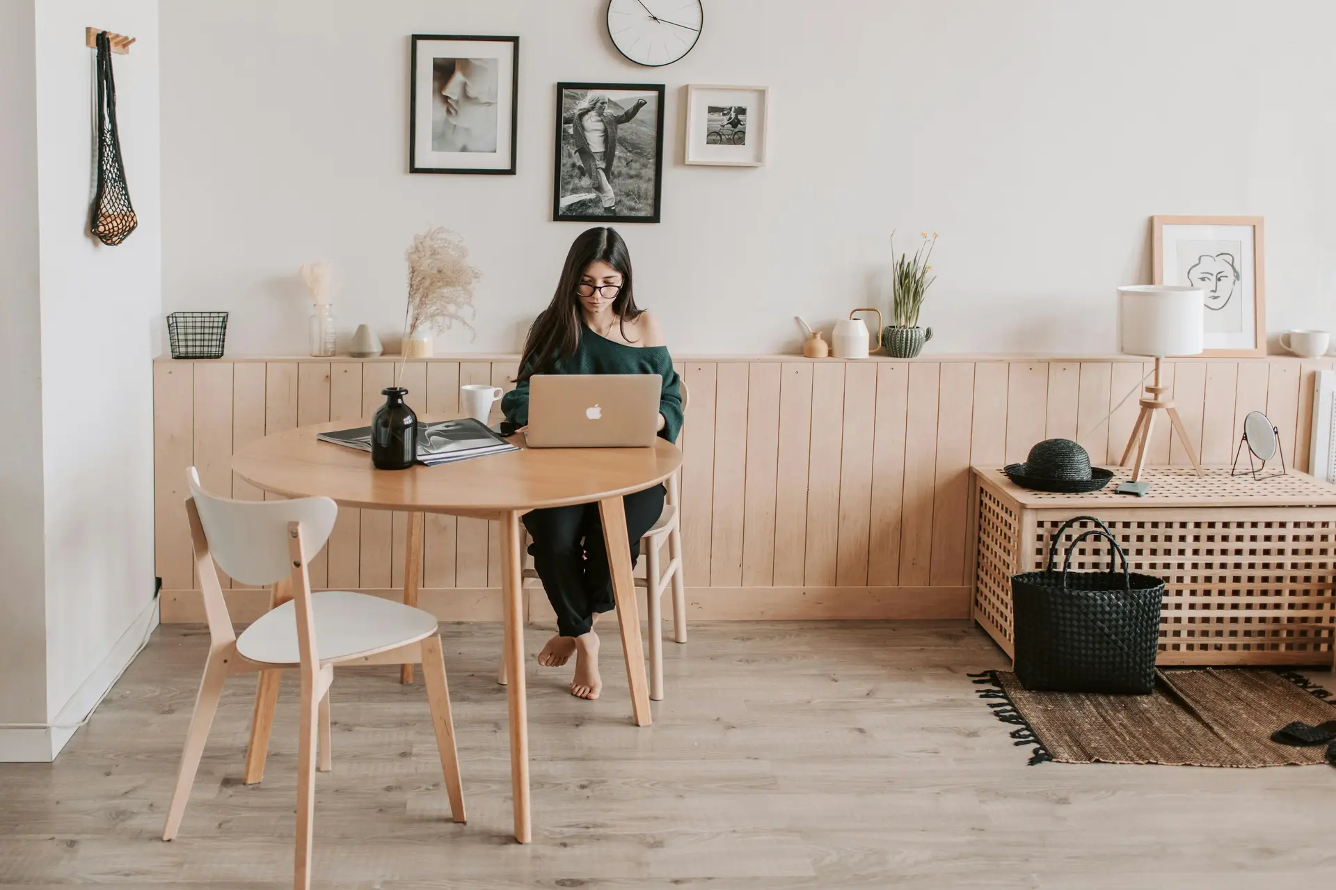 A bright Nordic living room featuring a calming wood and white combo, with a round wooden table, light chairs, and a woman working on her laptop. Soft wall art, simple decor pieces, and pale flooring create an airy, versatile space with a relaxed, modern feel.