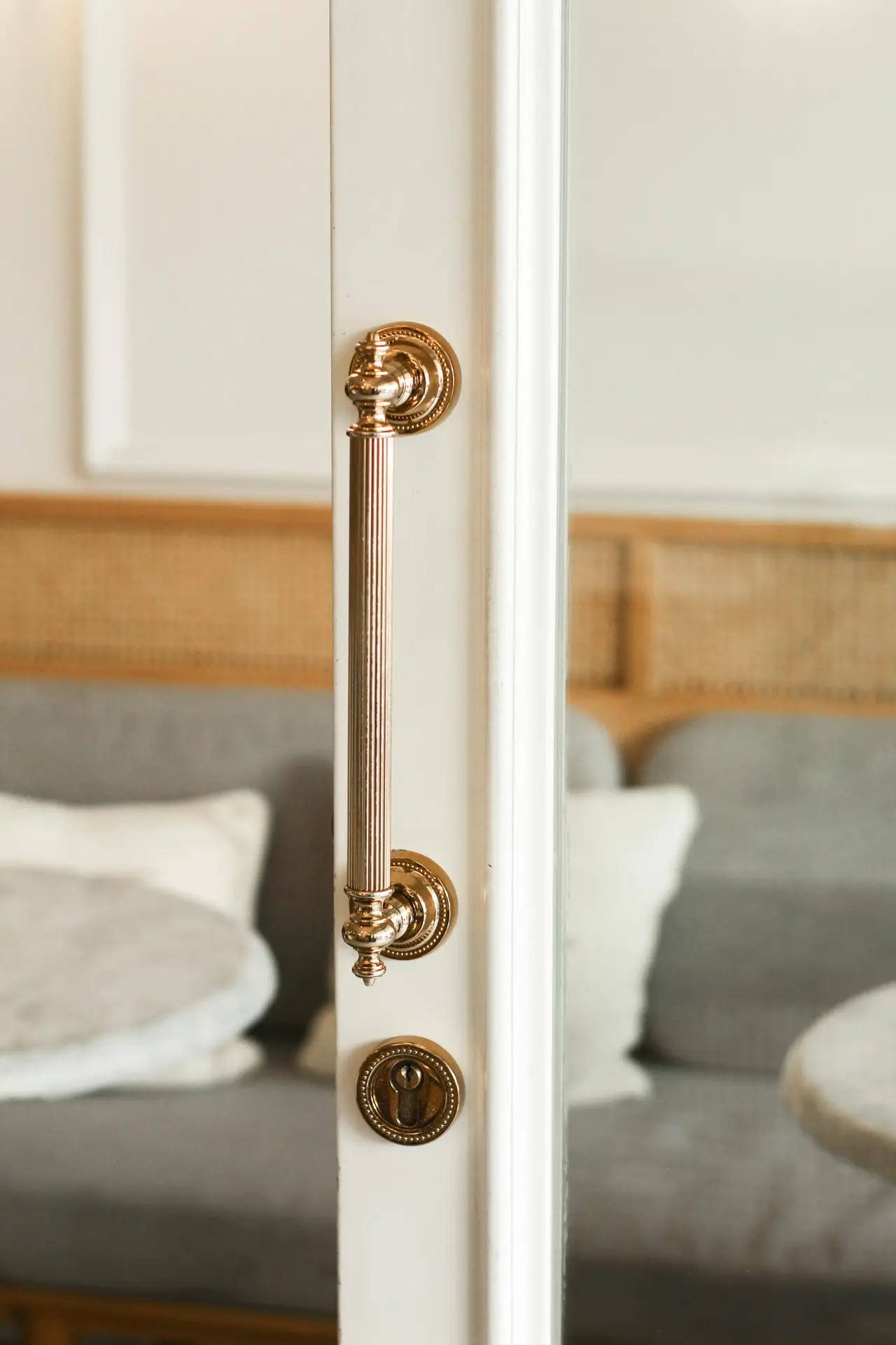 A close-up view of a polished brass door handle and lock on a white framed door, adding cohesive metal finishes to a calm setting styled with small living room decor. In the softly blurred background, a gray sofa with light pillows and woven accents creates a warm, inviting atmosphere that pairs naturally with the refined metallic details.