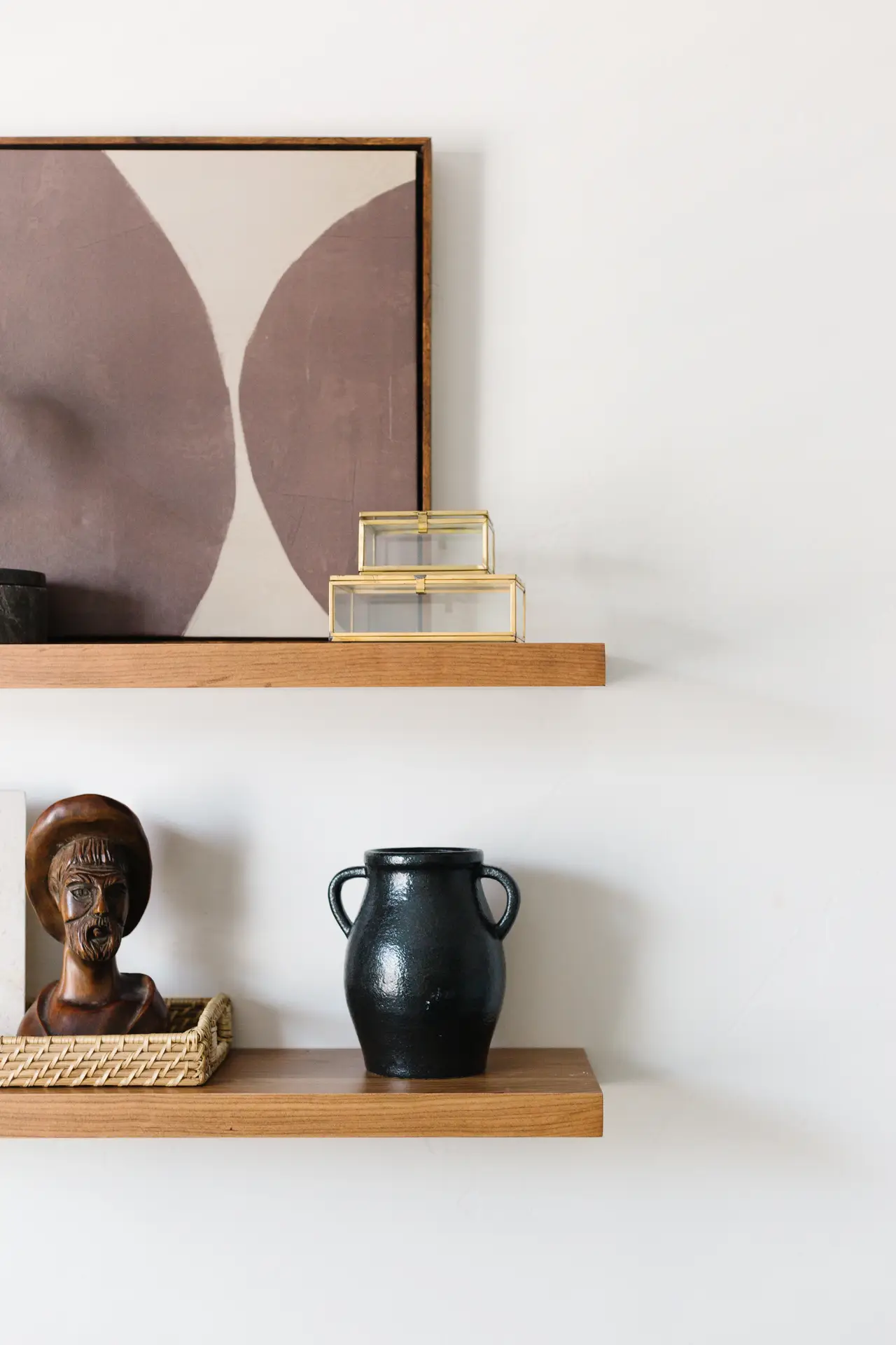 Two wooden floating shelves styled with small living room decor items, including abstract wall art, a black ceramic vase, gold glass boxes, and a carved wooden bust in a woven tray. The arrangement creates a modern and artistic display on a white wall.