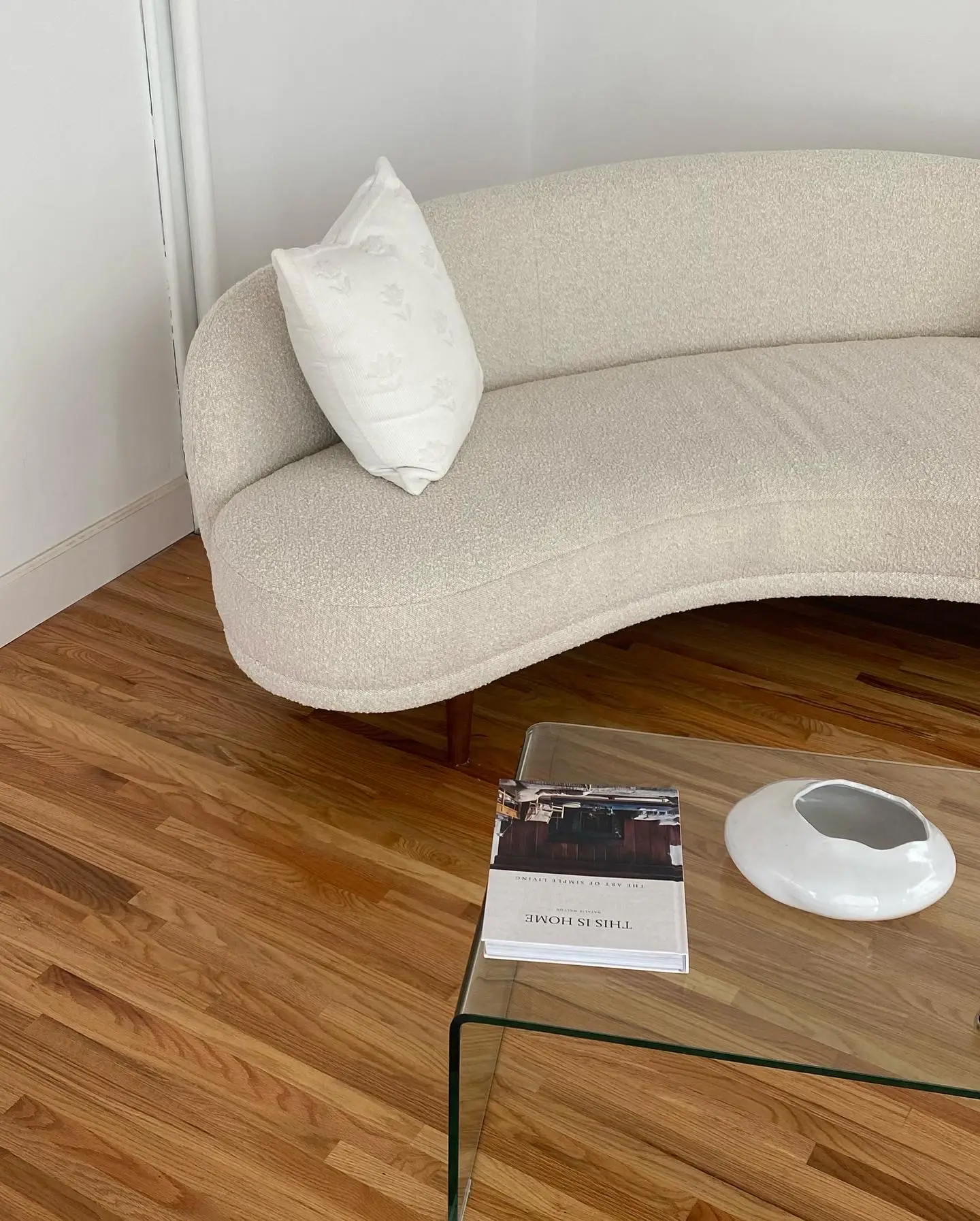 A cozy seating nook styled with small living room decor, featuring a curved cream sofa paired with a clear glass furniture piece acting as a minimalist coffee table. A design book and sculptural white bowl rest on top, while warm wood flooring adds contrast and makes the simple setup adaptable to many interiors.