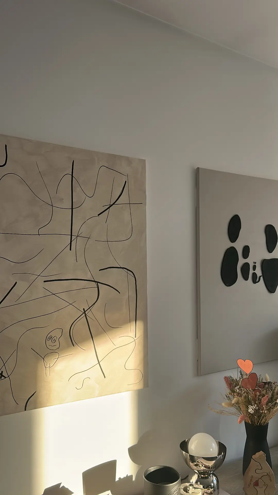 A bright corner styled with small living room decor, featuring two pieces of large-scale artwork on a neutral wall-one with abstract black linework and the other with minimal black shapes. Soft sunlight falls across the display, highlighting a mix of modern accents below, including a metallic lamp, a vase of dried flowers, and a few decorative objects.