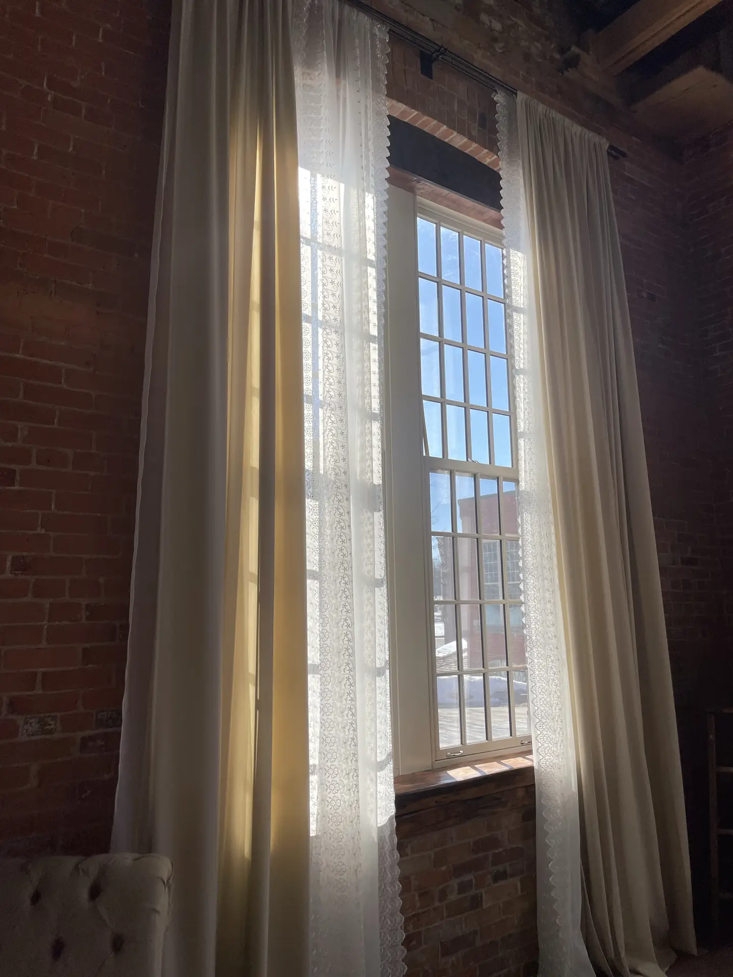 Tall windows framed by exposed brick walls are dressed with minimal curtain layers, combining soft cream panels and delicate sheer fabric to filter the sunlight. The setup adds warmth and height, making it a versatile example of small living room decor that brightens the space while keeping the look airy and understated.