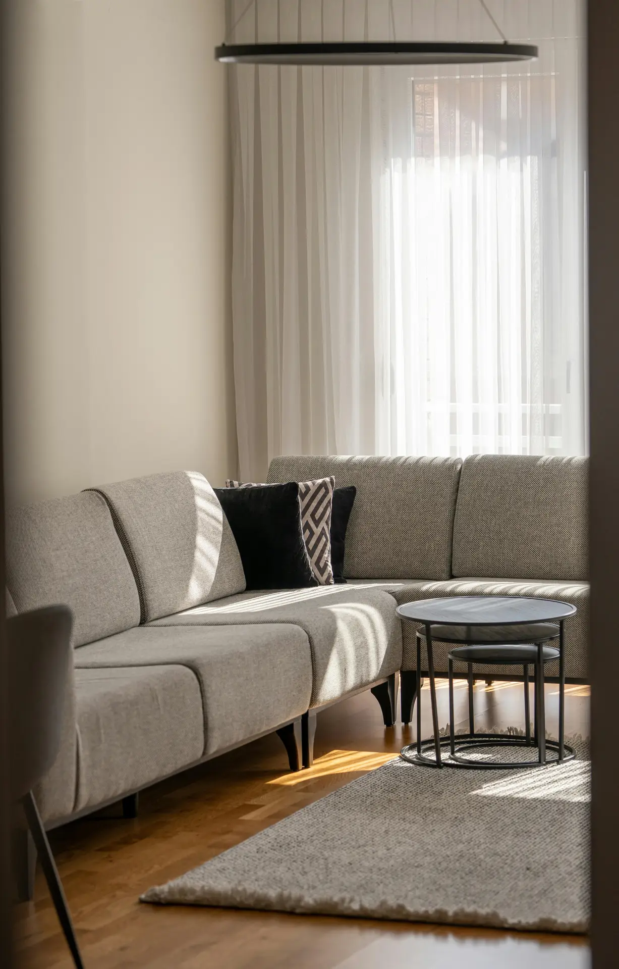 A cozy seating corner styled with small living room decor, showcasing a light gray sectional sofa paired with patterned and velvet throw pillows. Soft daylight filters through sheer curtains, brightening the space, while sleek black nesting tables sit on a textured rug, adding both function and a clean, modern touch.