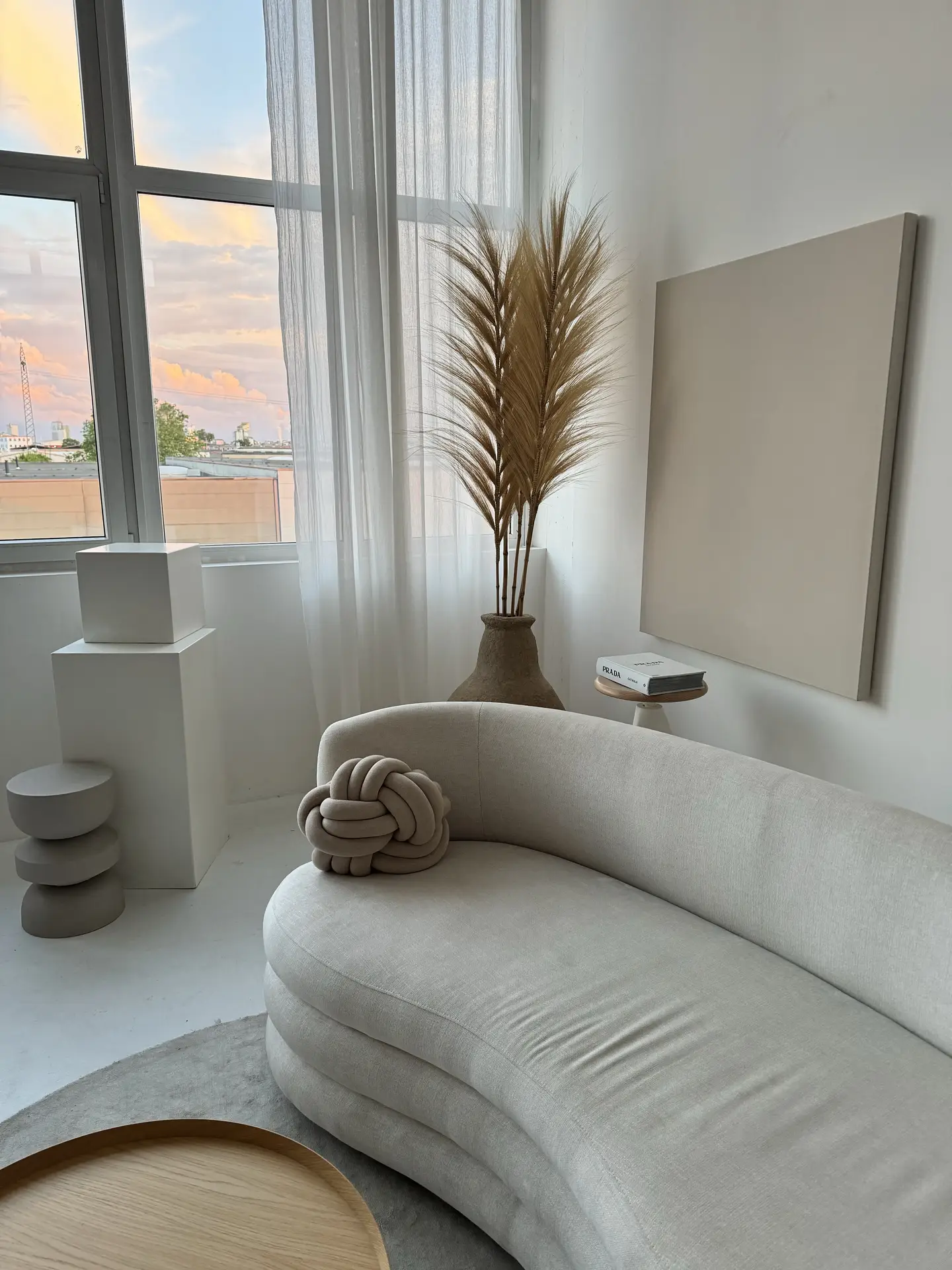 A soft, curved sofa in muted beige anchors this scene, showcasing small living room decor styled in neutral colors. Sheer curtains filter the sunset light, highlighting a sculptural vase with tall pampas stems, minimal wall art, and a round knot cushion. Clean lines, soft tones, and simple shapes create a calm and airy atmosphere.