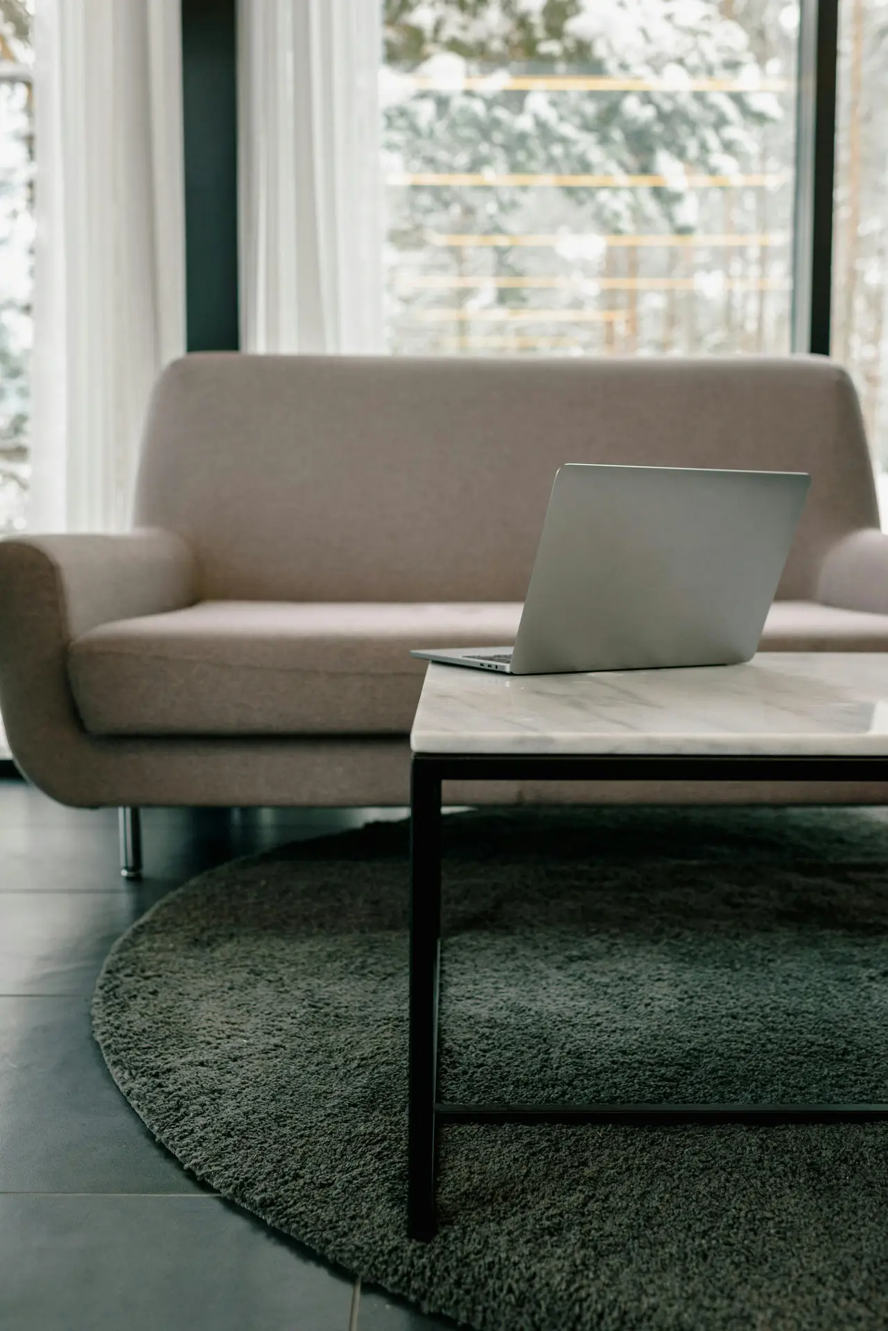 A soft, modern setup features a compact sofa facing a marble-top coffee table, with a laptop placed on it. Beneath the table lies a textured round rug that helps define the seating zone, making the scene a clean and versatile example of small living room decor that balances comfort and functionality.