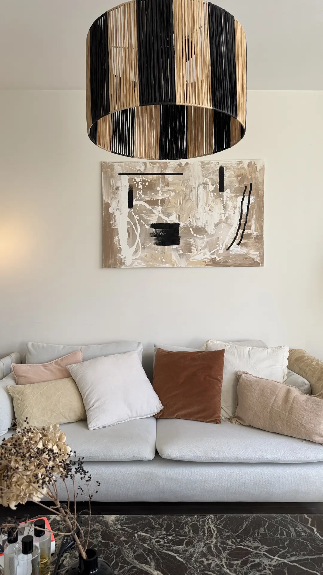 A cozy seating area styled with small living room decor features a soft, light-colored sofa layered with mixed neutral pillows, paired with a bold abstract artwork on the wall. Above it hangs a statement lighting fixture made of woven black and natural fibers, adding height and visual interest. A dark marble coffee table and dried floral accents complete the warm, versatile setup.