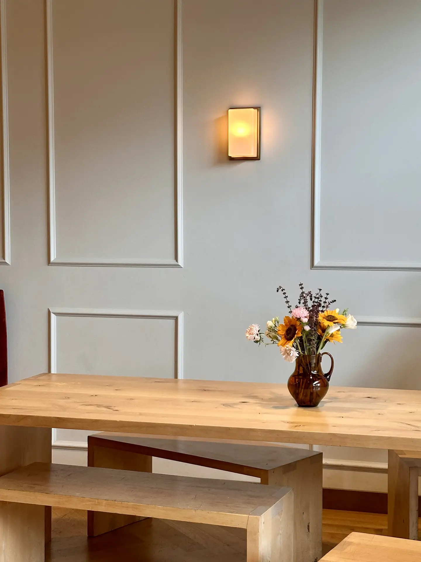 A soft, neutral corner styled with small living room decor, featuring a simple wooden table and bench set topped with a brown vase of mixed flowers. Above it, warm wall-mounted lighting glows against paneled walls, creating a cozy yet adaptable atmosphere for different interior styles.