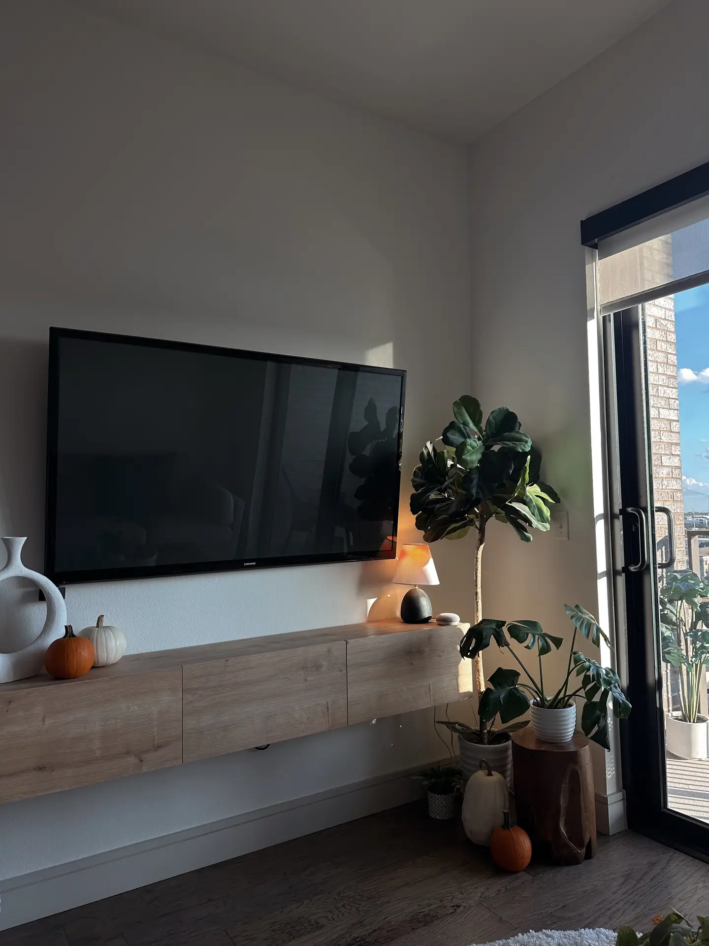 A cozy corner setup featuring a wall-mounted TV above a light wood floating console styled with small pumpkins and modern vases. To the right, a cluster of potted plants sits on natural wood stools near a sliding glass door, bringing in daylight and greenery. This arrangement highlights small living room decor that mixes warm accents, simple surfaces, and a practical wall-mounted tv for a clean, open feel.