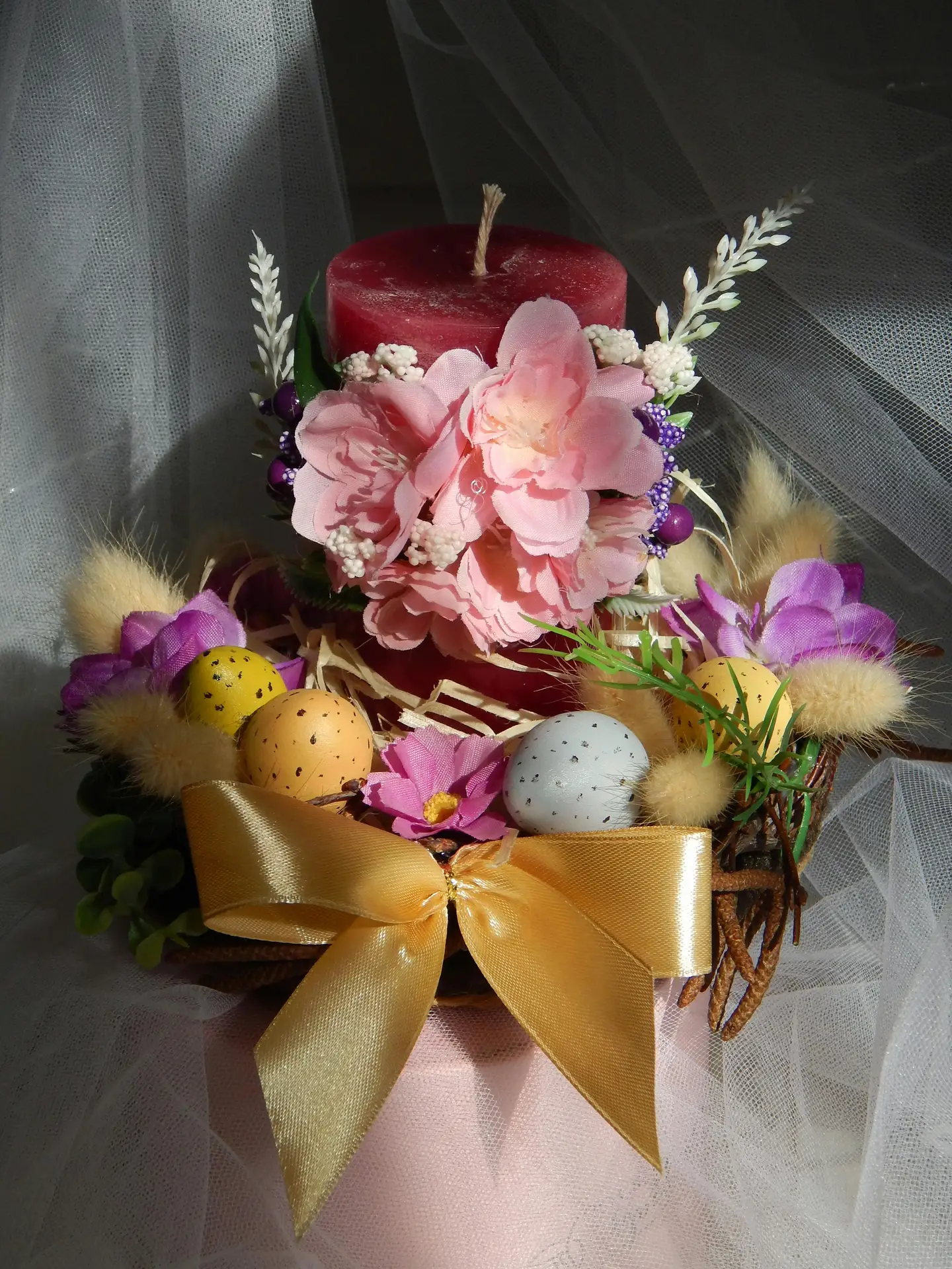 Easter decor ideas featuring an arrangement with candle, flowers and eggs, showing a deep red pillar candle decorated with soft pink blossoms, surrounded by speckled decorative eggs, natural twigs, delicate greenery, and a golden ribbon, creating a layered spring centerpiece with a cozy, festive feel.