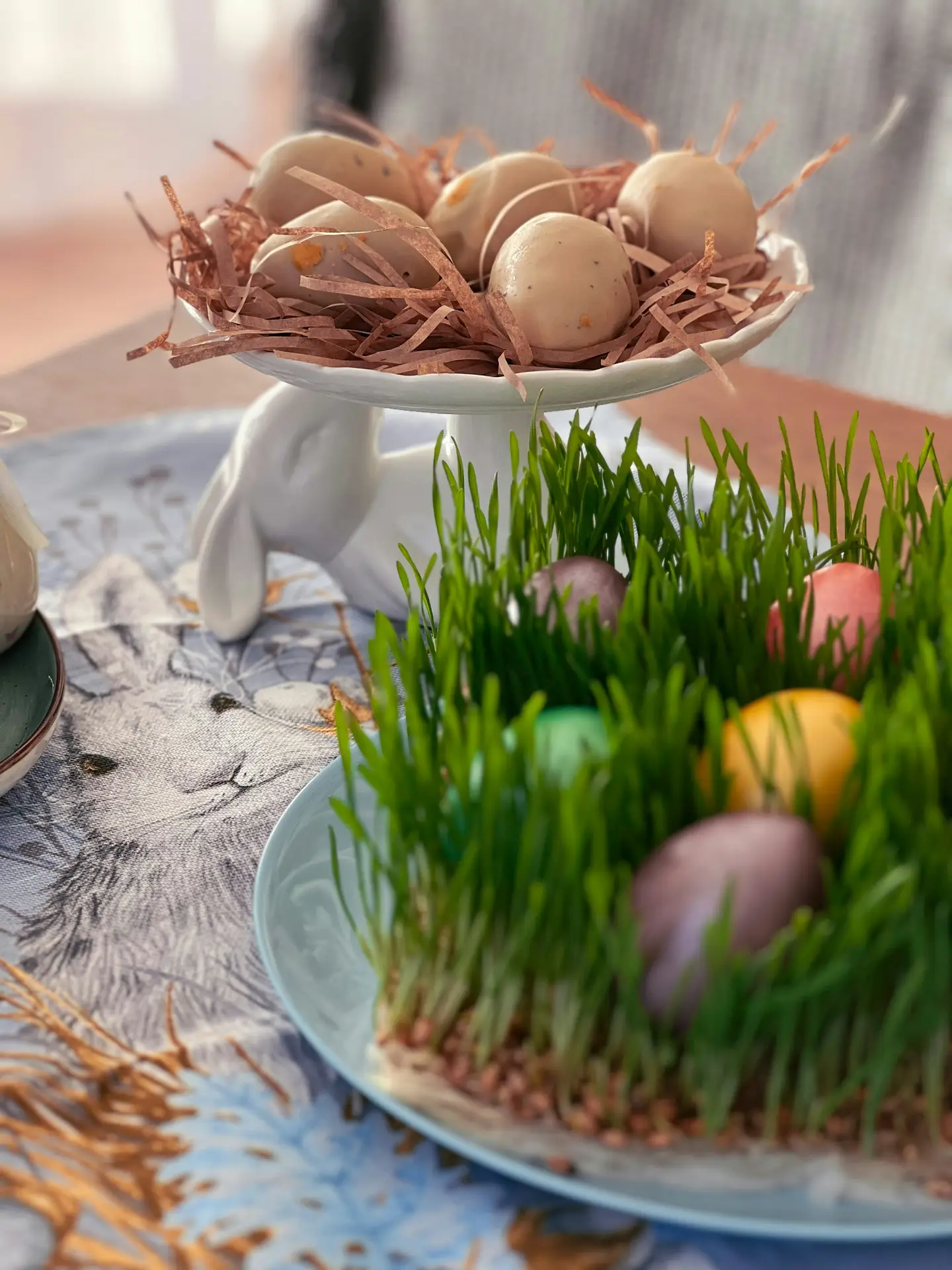 Easter decor ideas showing a bunny tiered egg stand holding neutral-toned eggs nestled in paper straw, paired with a plate of fresh green grass and colorful eggs on a softly styled spring table setting.