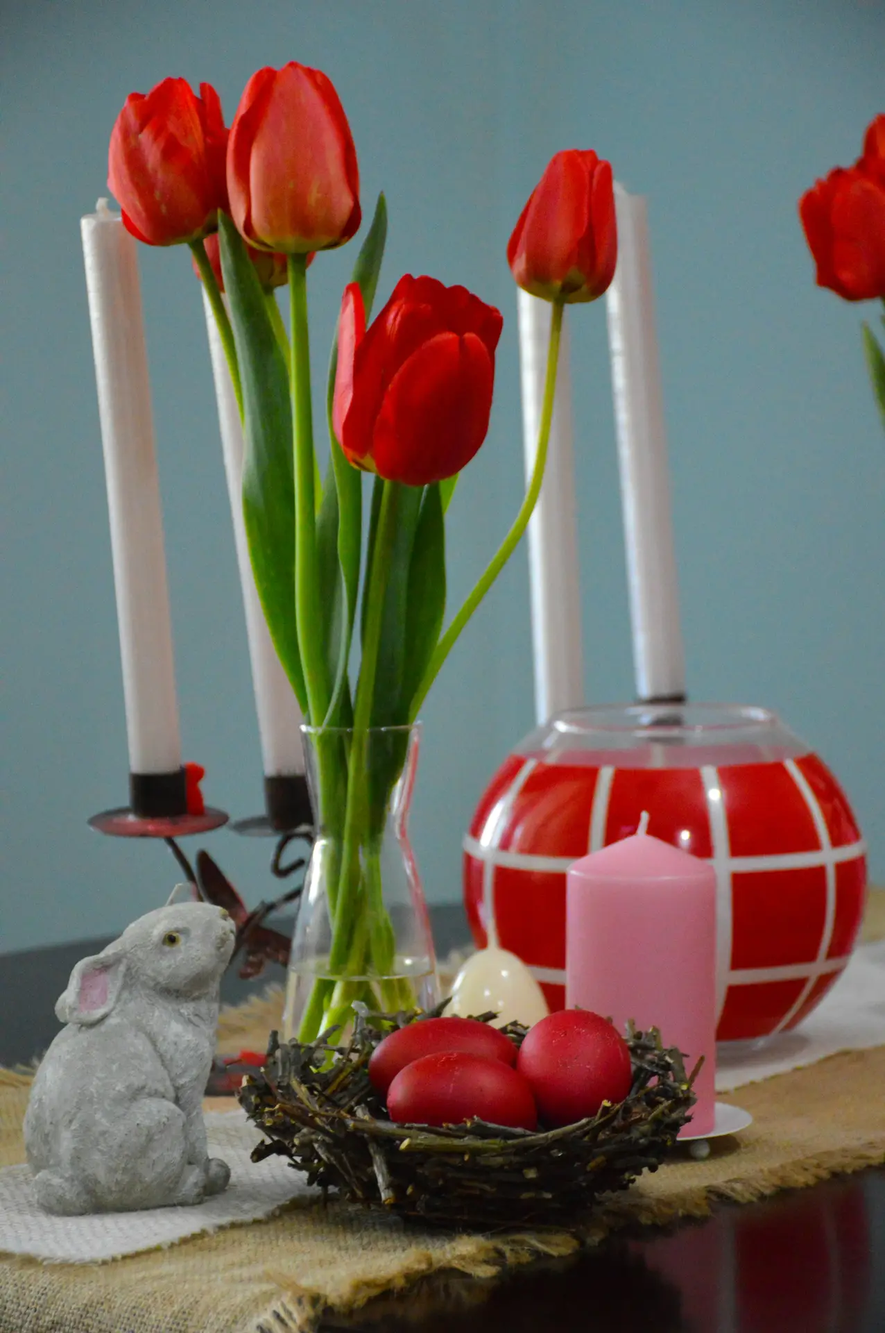Easter decor ideas featuring an easter nest with rabbit and tulips, showing a small woven nest filled with red eggs, a stone bunny figurine, fresh red tulips in a glass vase, and soft candle accents arranged on a rustic tabletop.