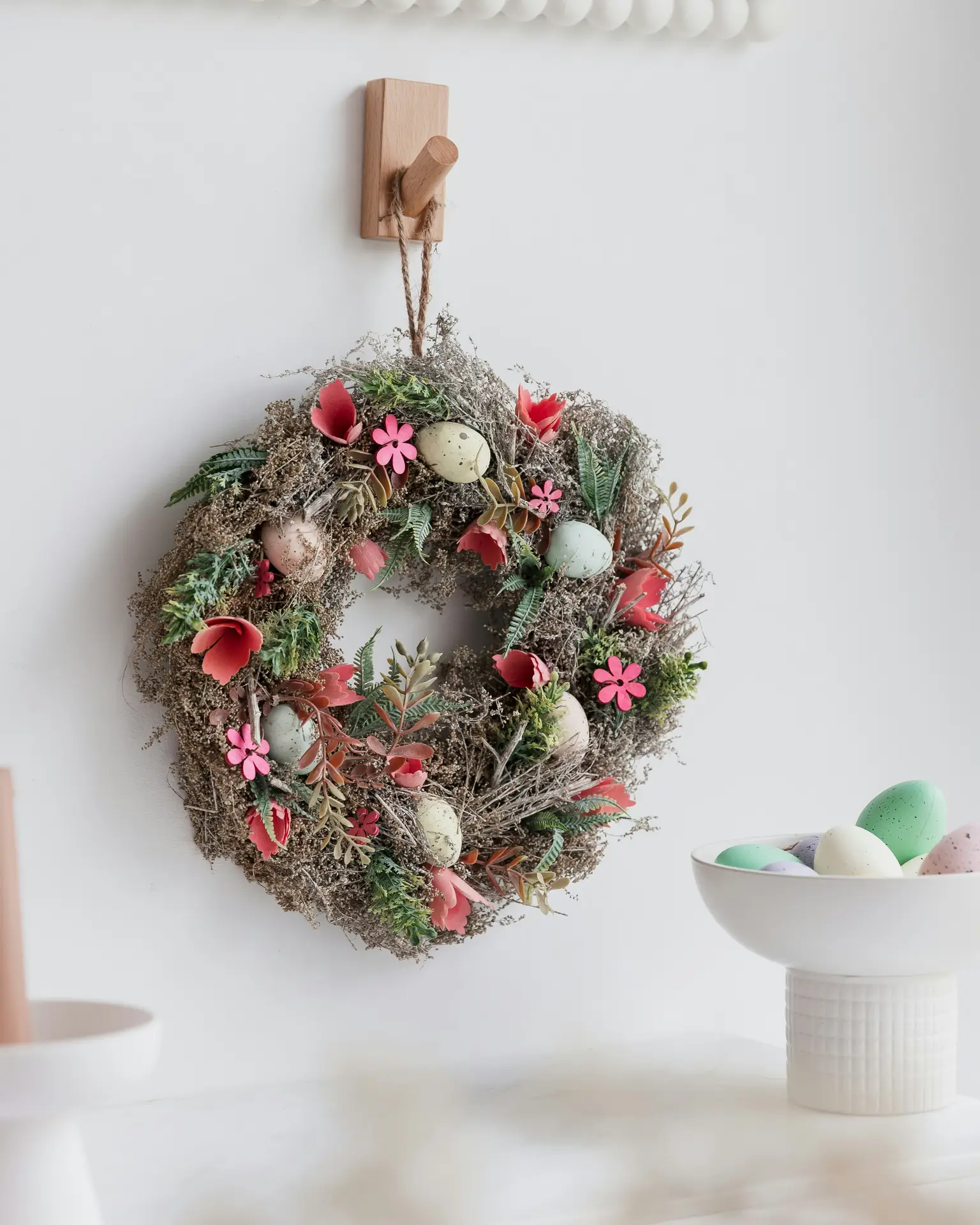 Easter decor ideas showing a handmade easter wreath hanging on a wall, styled with dried greenery, small pastel eggs, and soft floral accents, paired with a minimal shelf and a bowl of decorative eggs for a calm seasonal look.