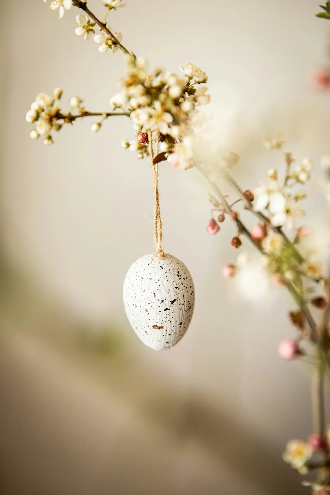 Easter decor ideas showcasing an egg hanger variation, with a speckled white decorative egg suspended by natural twine from a delicate flowering branch, creating a soft and airy spring-inspired accent against a neutral background.