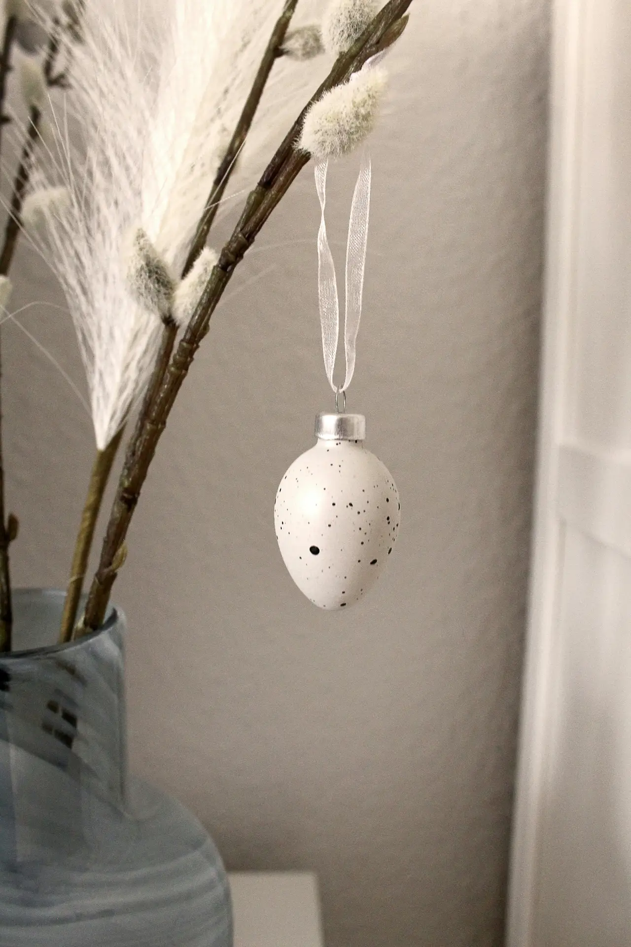 Easter decor ideas featuring a delicate egg hanger with a speckled white ornament suspended from a soft ribbon, styled on natural branches with fluffy catkins in a simple ceramic vase for a calm, minimalist seasonal display.