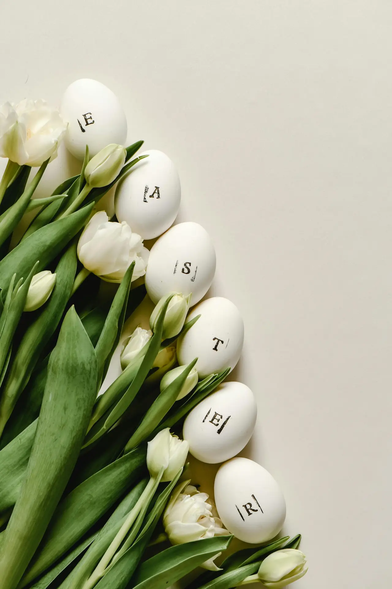 Easter decor ideas featuring painted eggs with subtle lettering arranged among fresh white tulips and green leaves on a light background, creating a soft and elegant spring-inspired composition.