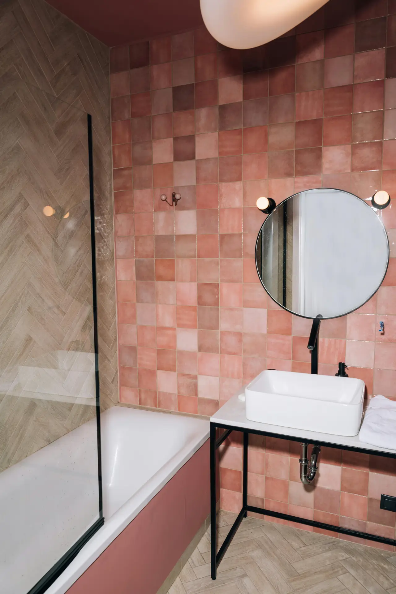 A modern bathroom with soft blush tiles covering the walls, paired with a minimalist vanity and a round mirror framed in black. A glass shower panel and light wood flooring balance the warm pink tones, creating an elegant yet playful look. This setup showcases girls bathroom ideas that use blush tiles or pastel walls to add color while keeping the space stylish and grown-up.